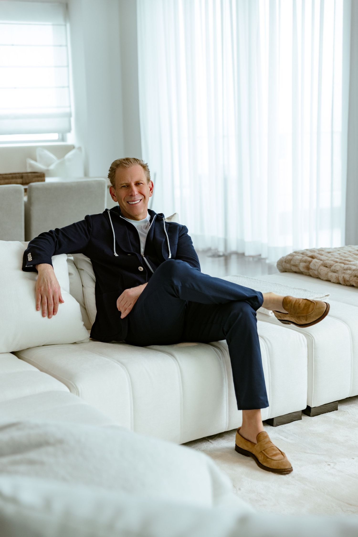 Professional portrait of Michael Liebowitz seated casually on a white sectional in a bright, contemporary living space.