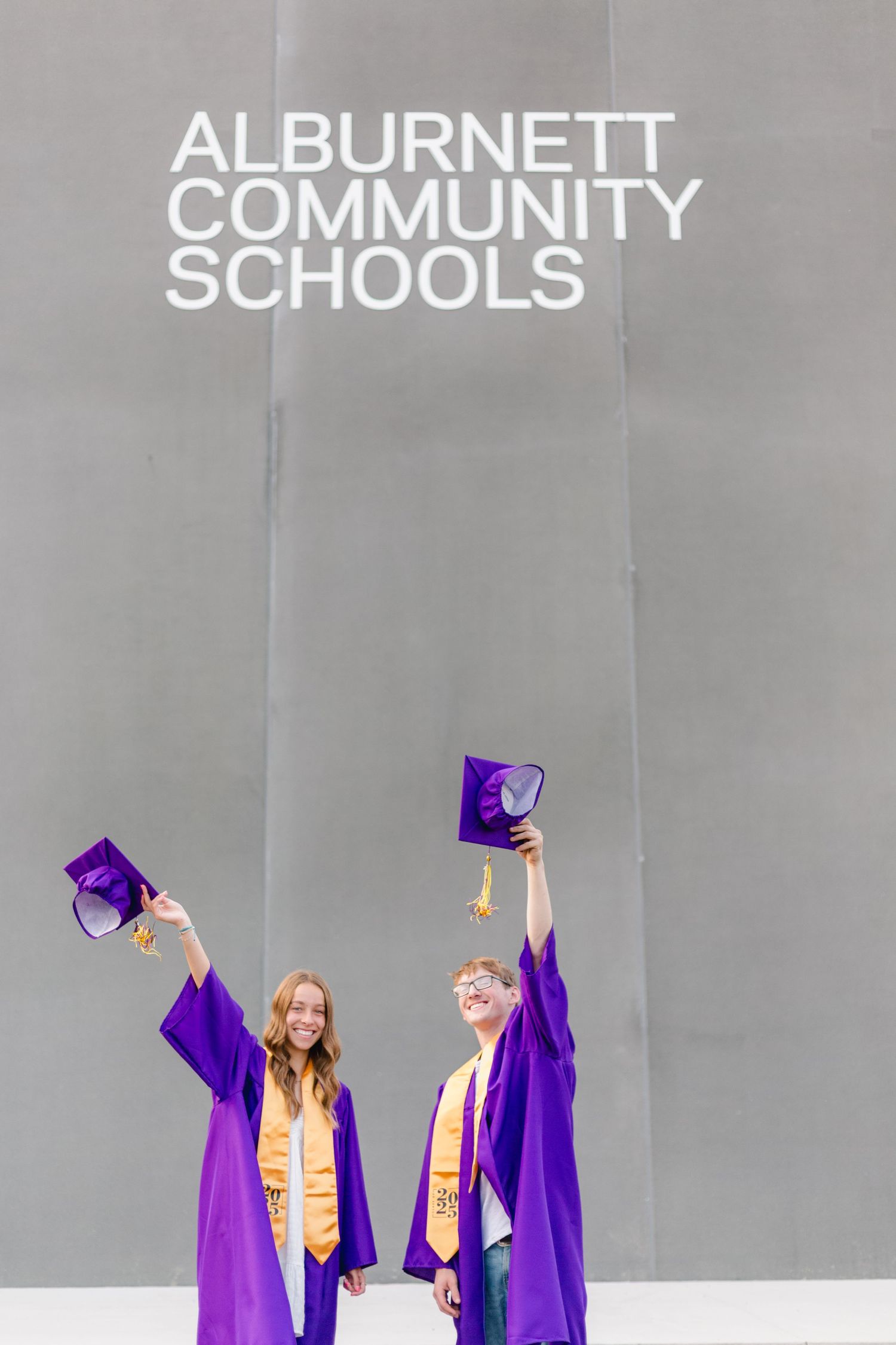 Students in purple graduation gowns celebrate together against a gray background at Alburnett Community Schools with Nichole Sieck Photo.