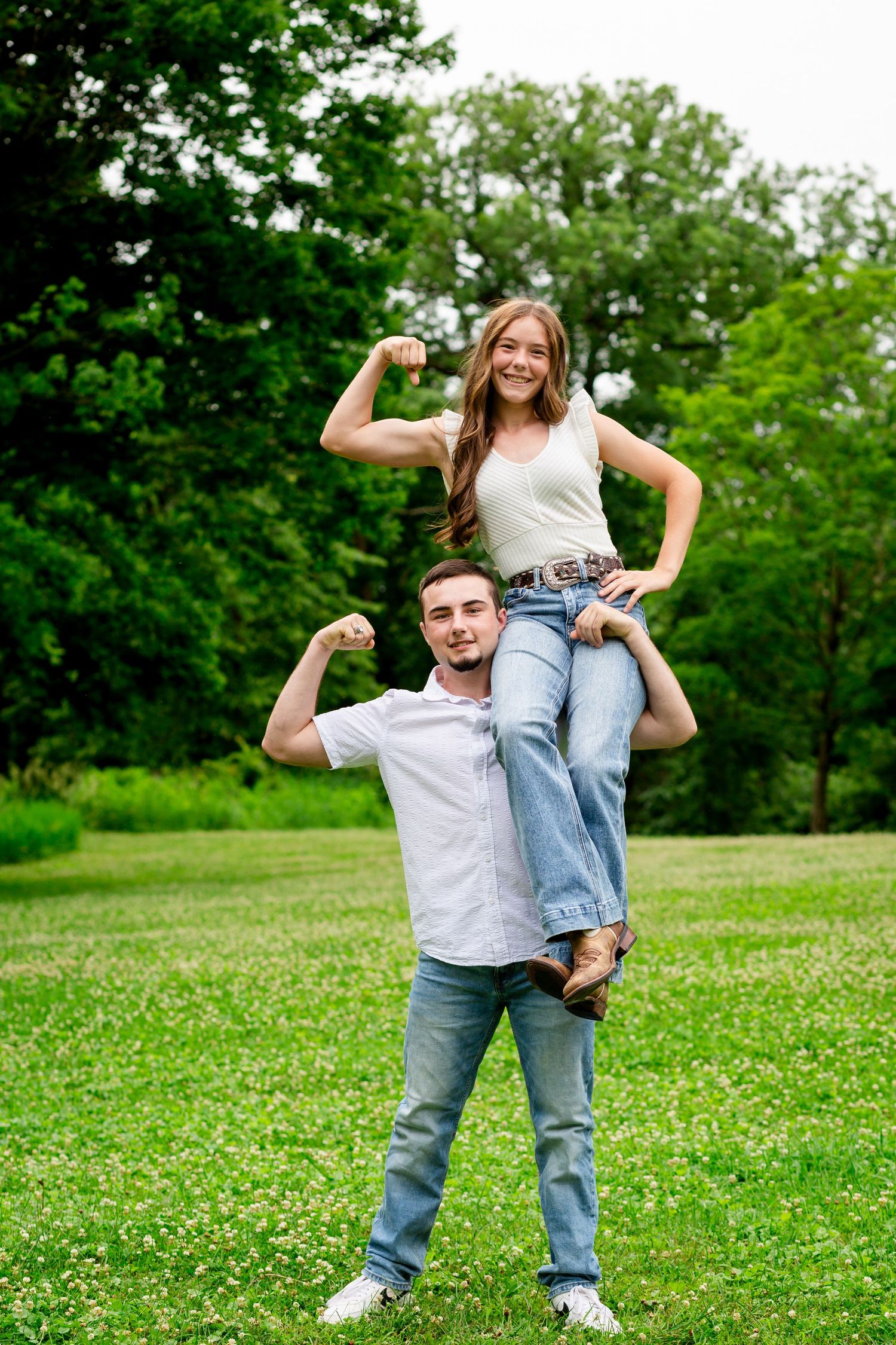 A young couple flexing their muscles outdoors, with the man carrying the woman on his shoulder in a green park.