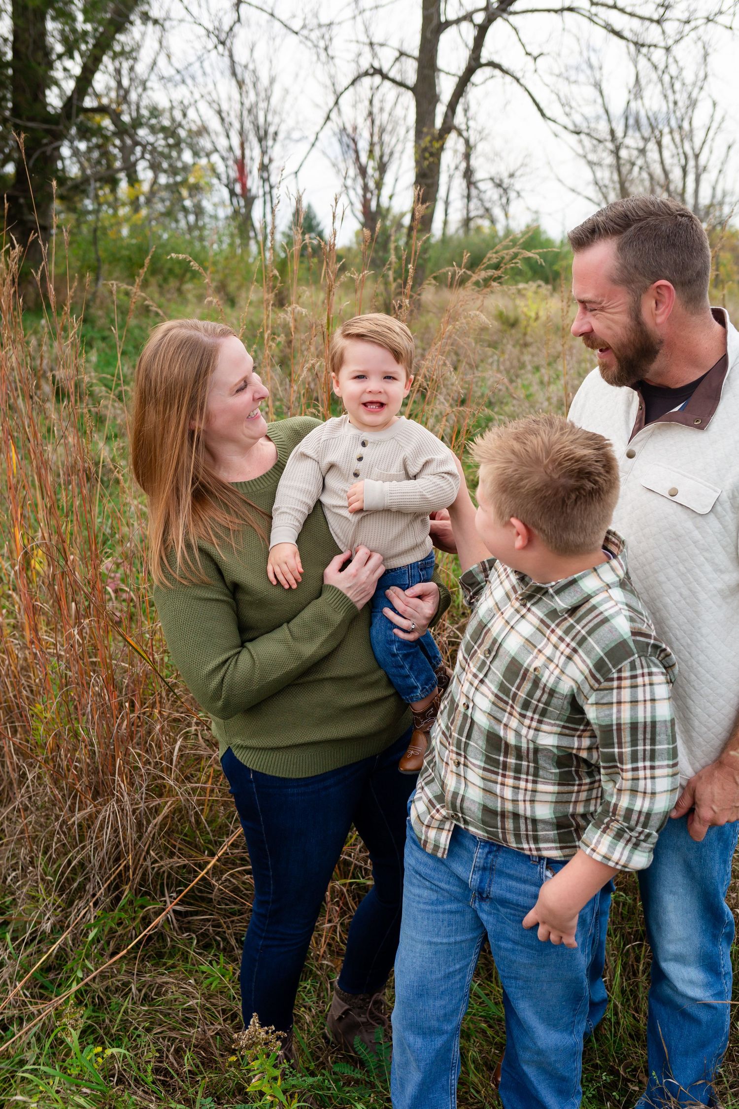 Family of four enjoying outdoor fall photo session with two young boys, surrounded by autumn foliage and tall grass.