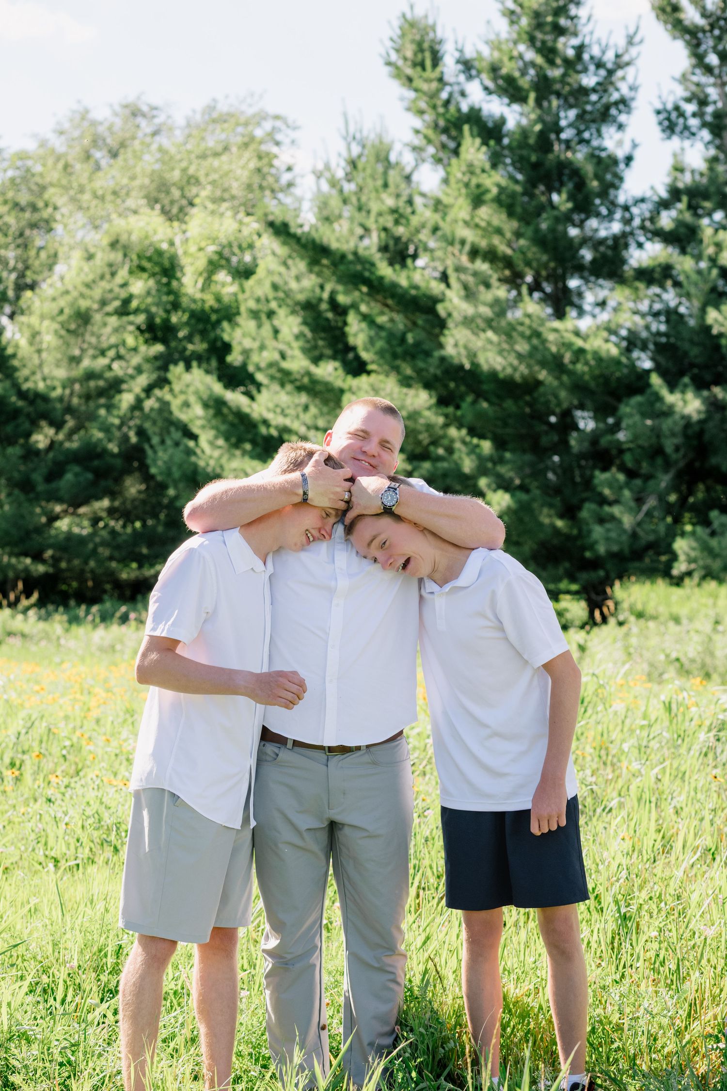 A smiling father playfully hugs his two teenage sons outdoors in a sunny green field.
