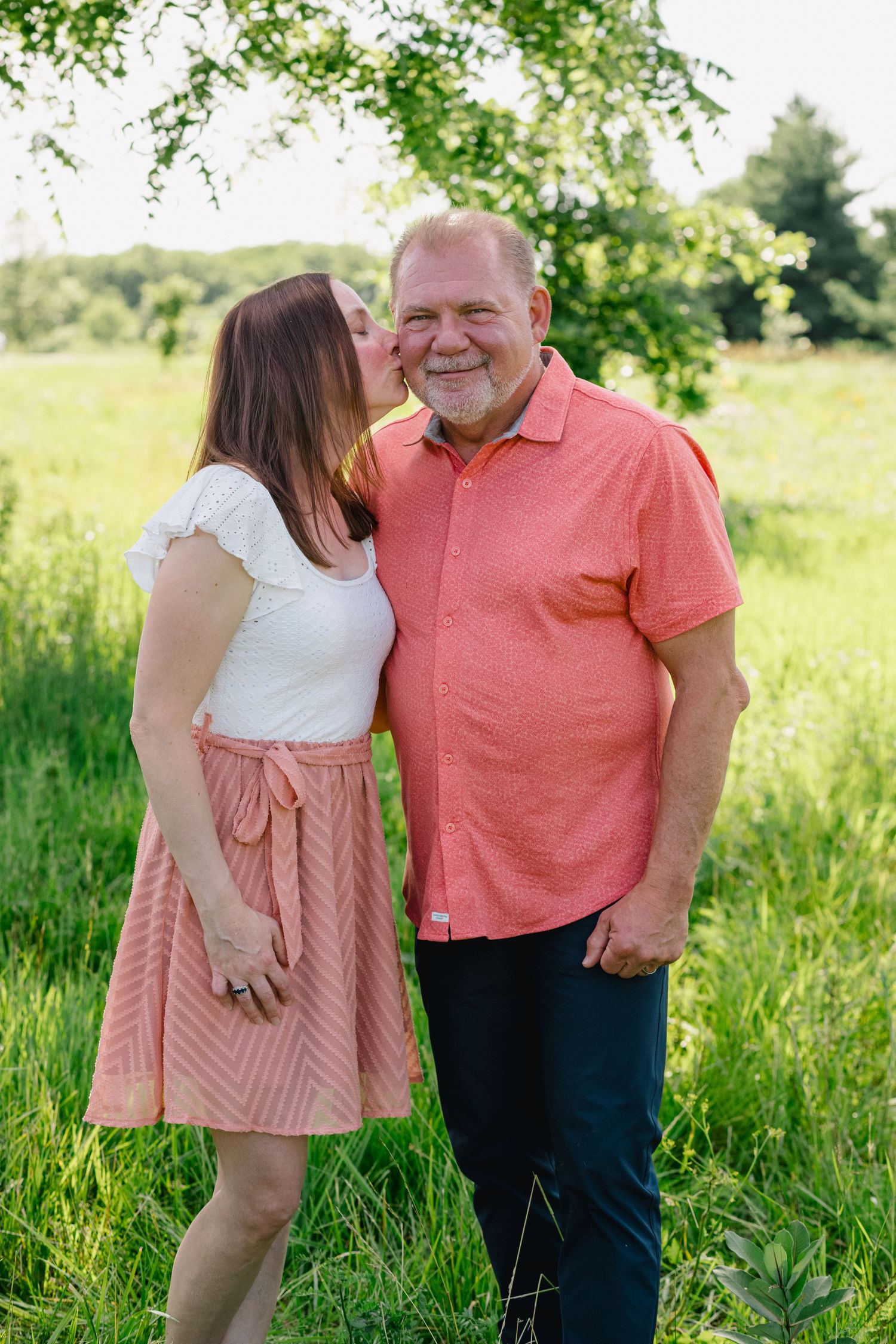 Couple posing outdoors in green field, woman kissing mans cheek, wearing coordinating pink outfits.