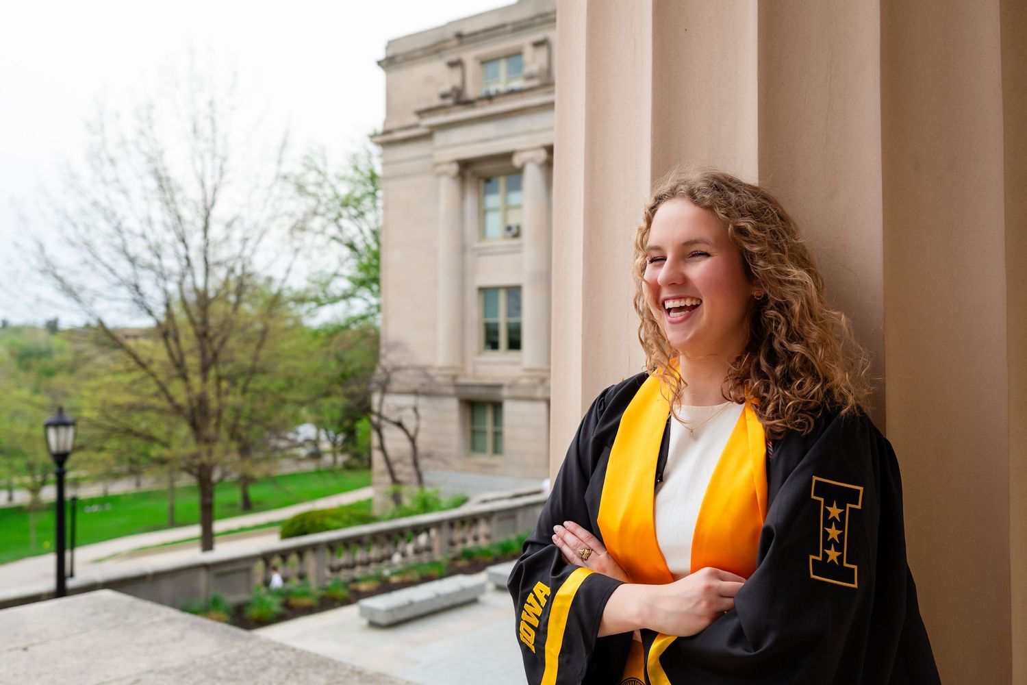 A smiling female graduate in black cap and gown with gold stole poses outdoors near a stone column on a college campus.