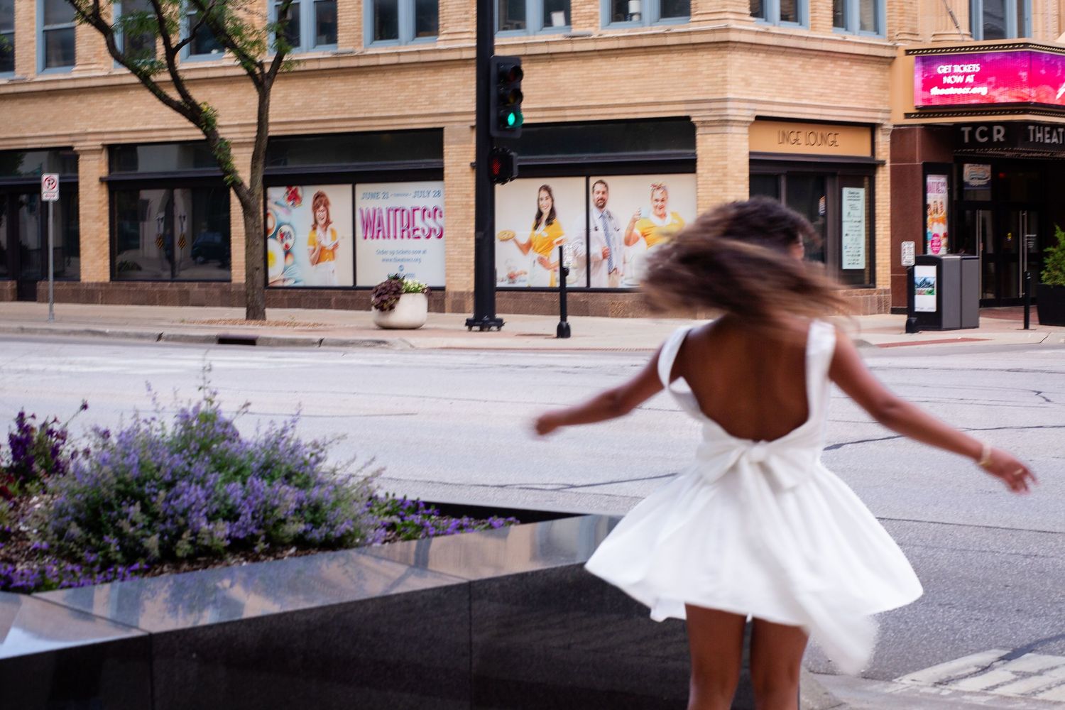 Young girl in white backless dress spinning outdoors near urban flower bed with Waitress musical poster in background.