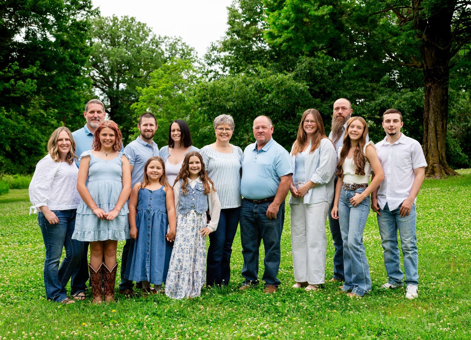 Large family group portrait smiling outdoors on green grass with lush trees in background on a summer day.