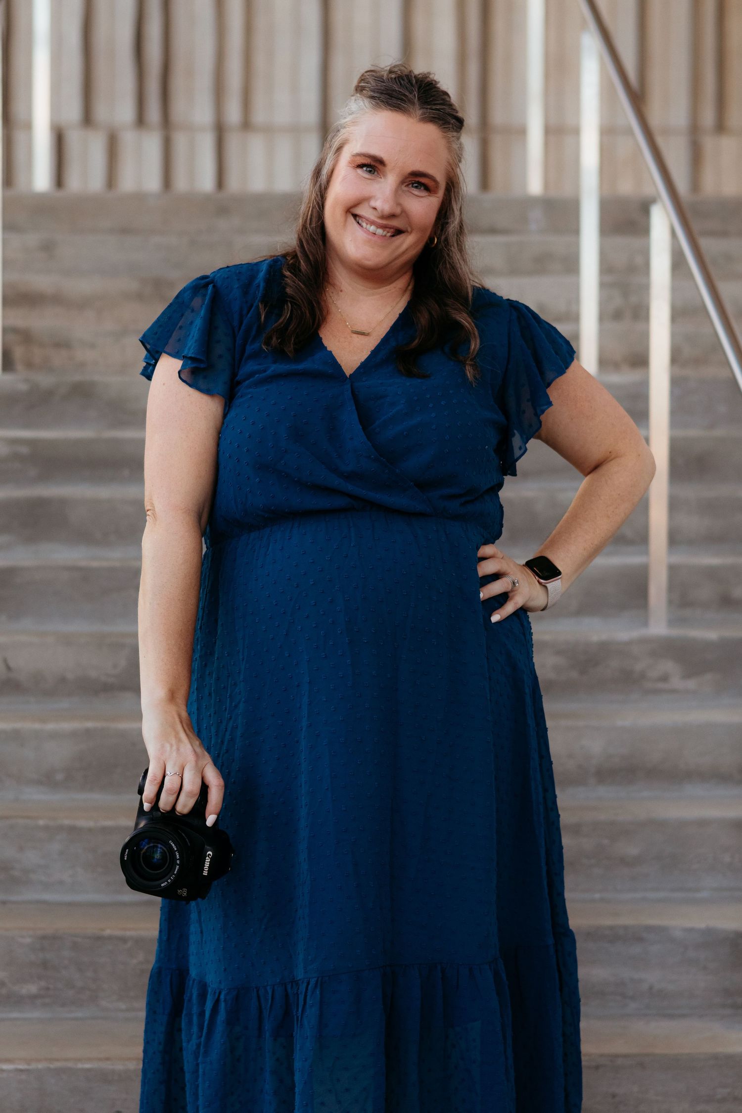 A photographer in a navy blue dress poses with a camera in a concrete architectural setting.