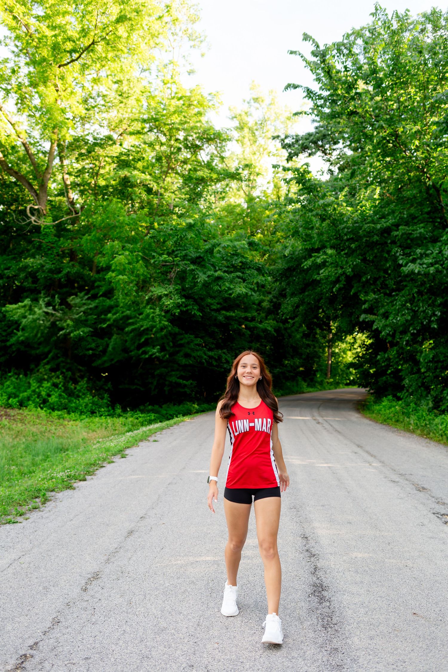 Young woman in red athletic tank and black shorts walks on a tree-lined road on a sunny day.