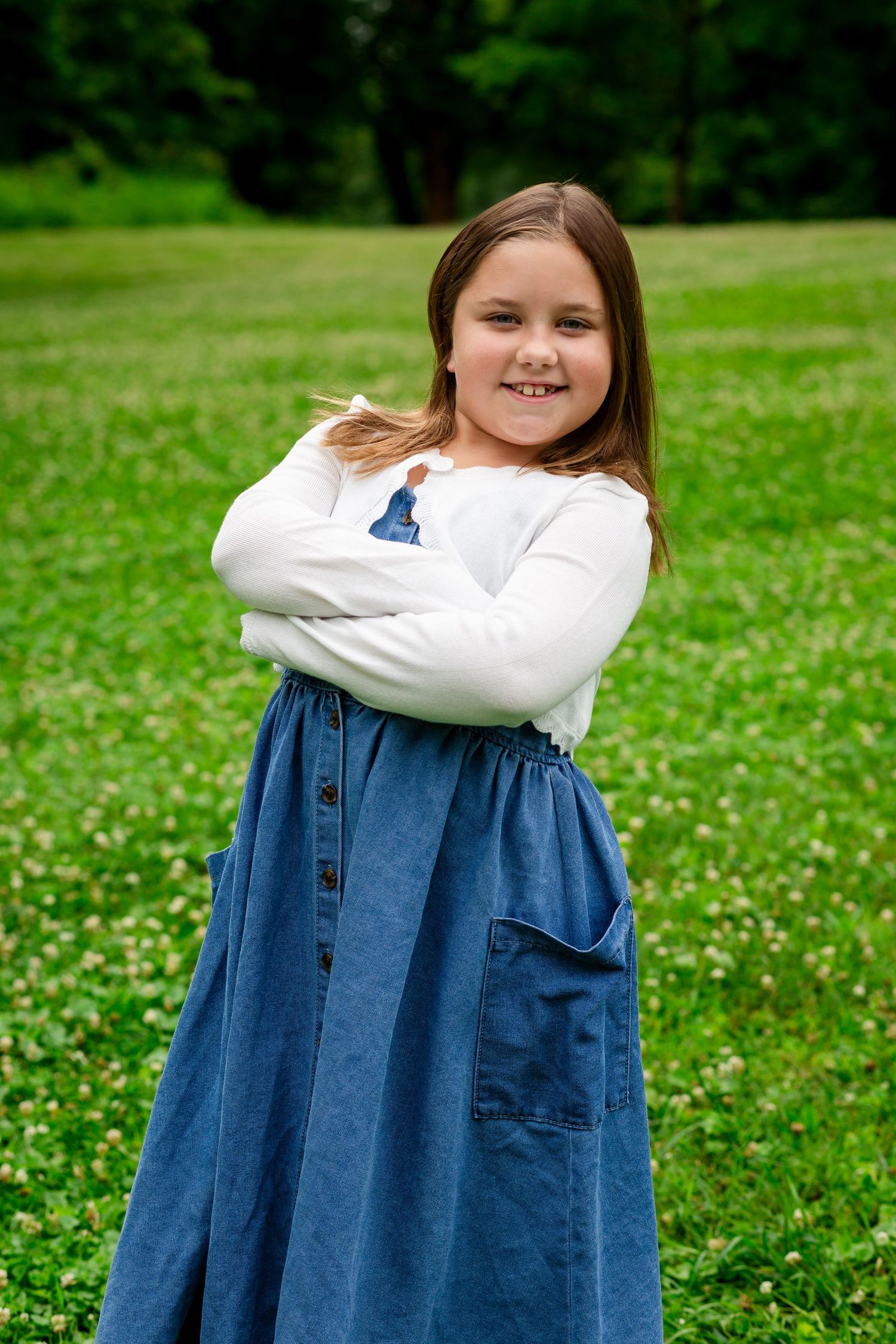 Young girl in denim skirt and white top posing with arms crossed in a green field.