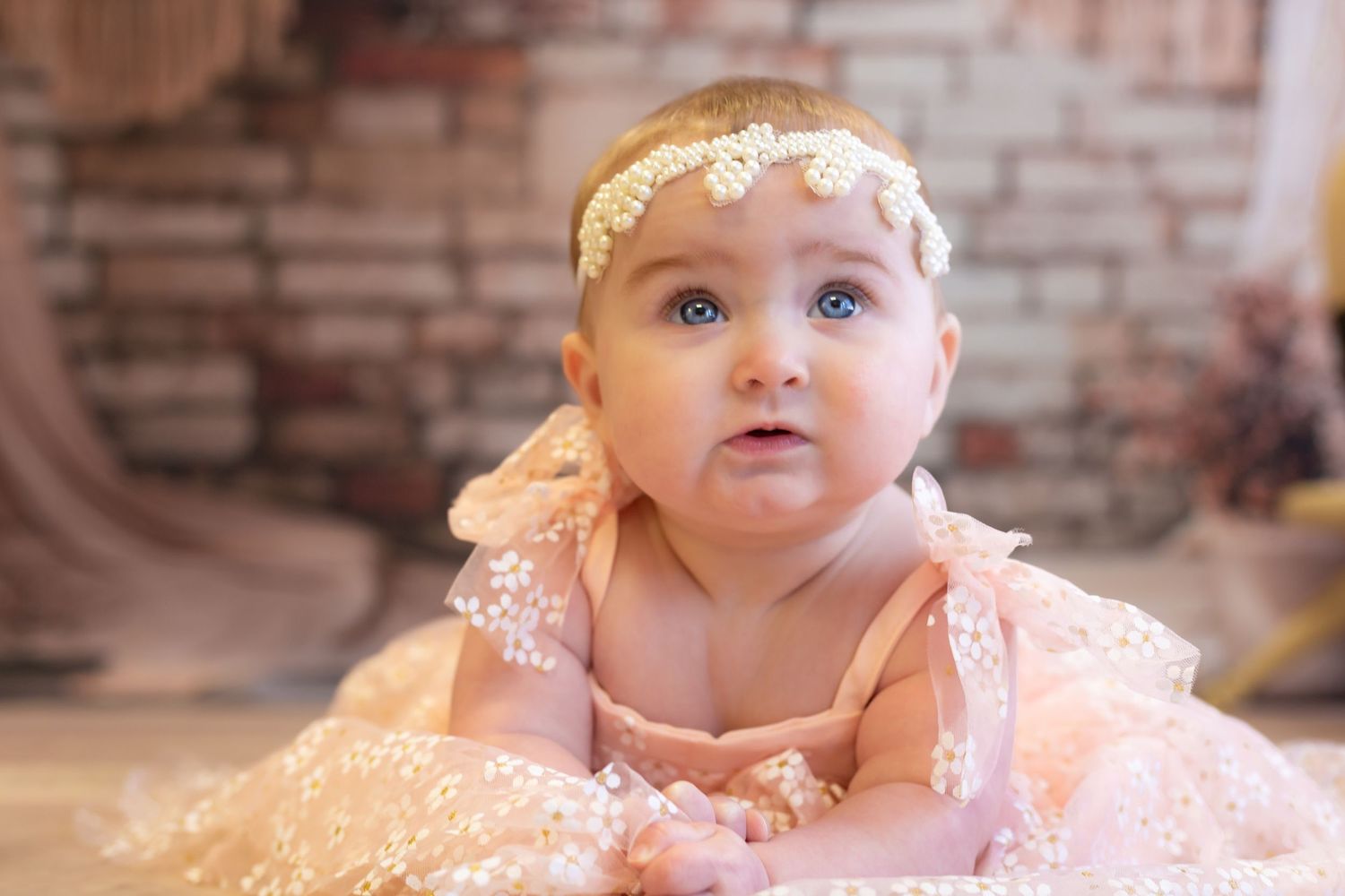 Sweet baby in a peach tulle dress and floral headband sits in front of brick wall in soft dreamy lighting.