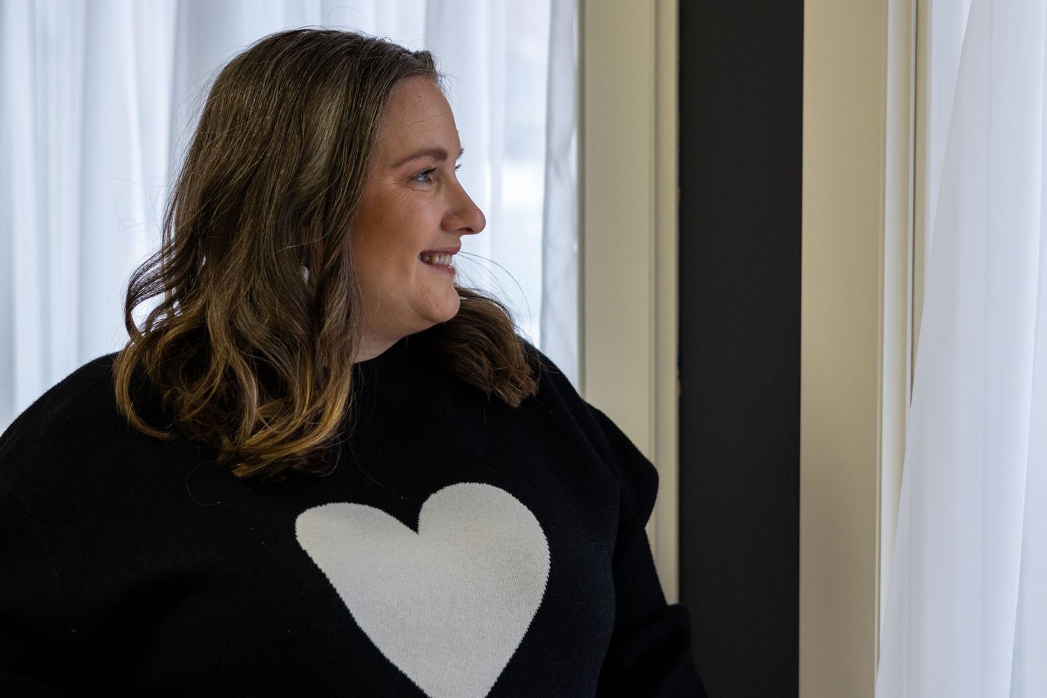 Nichole Sieck with Nichole Sieck Photography is in a black sweater with a white heart design laughs while looking towards a window.