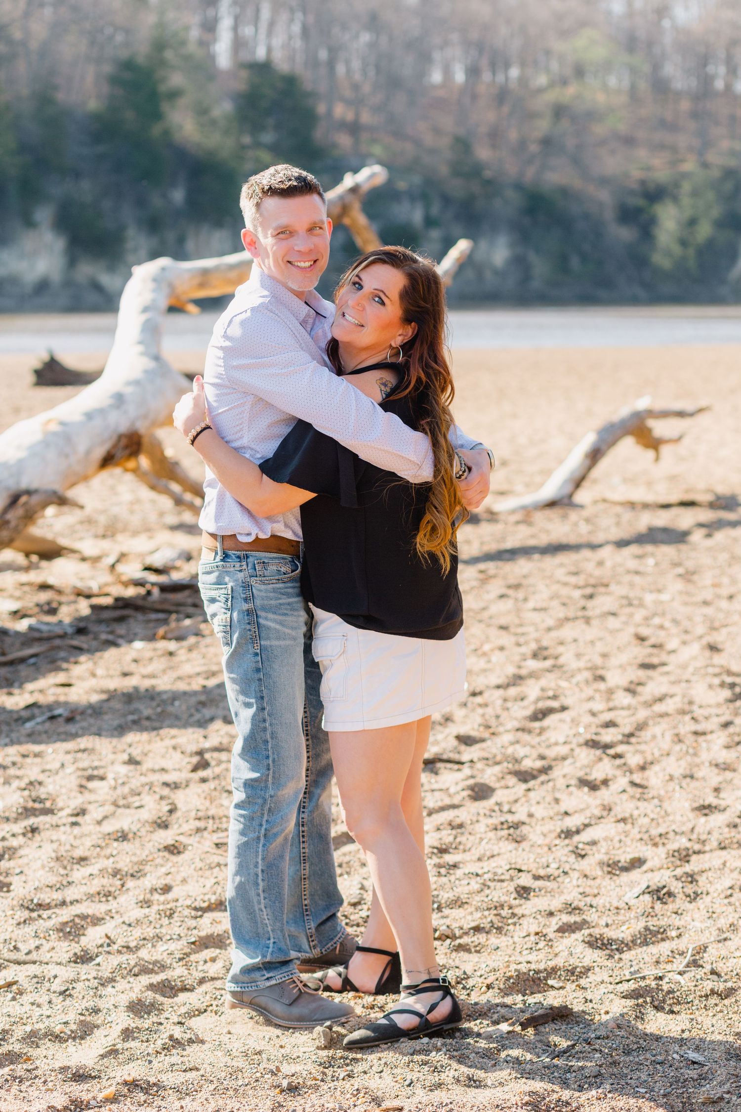 Happy couple embraces on a sunny beach with driftwood in the background on a beautiful autumn day.