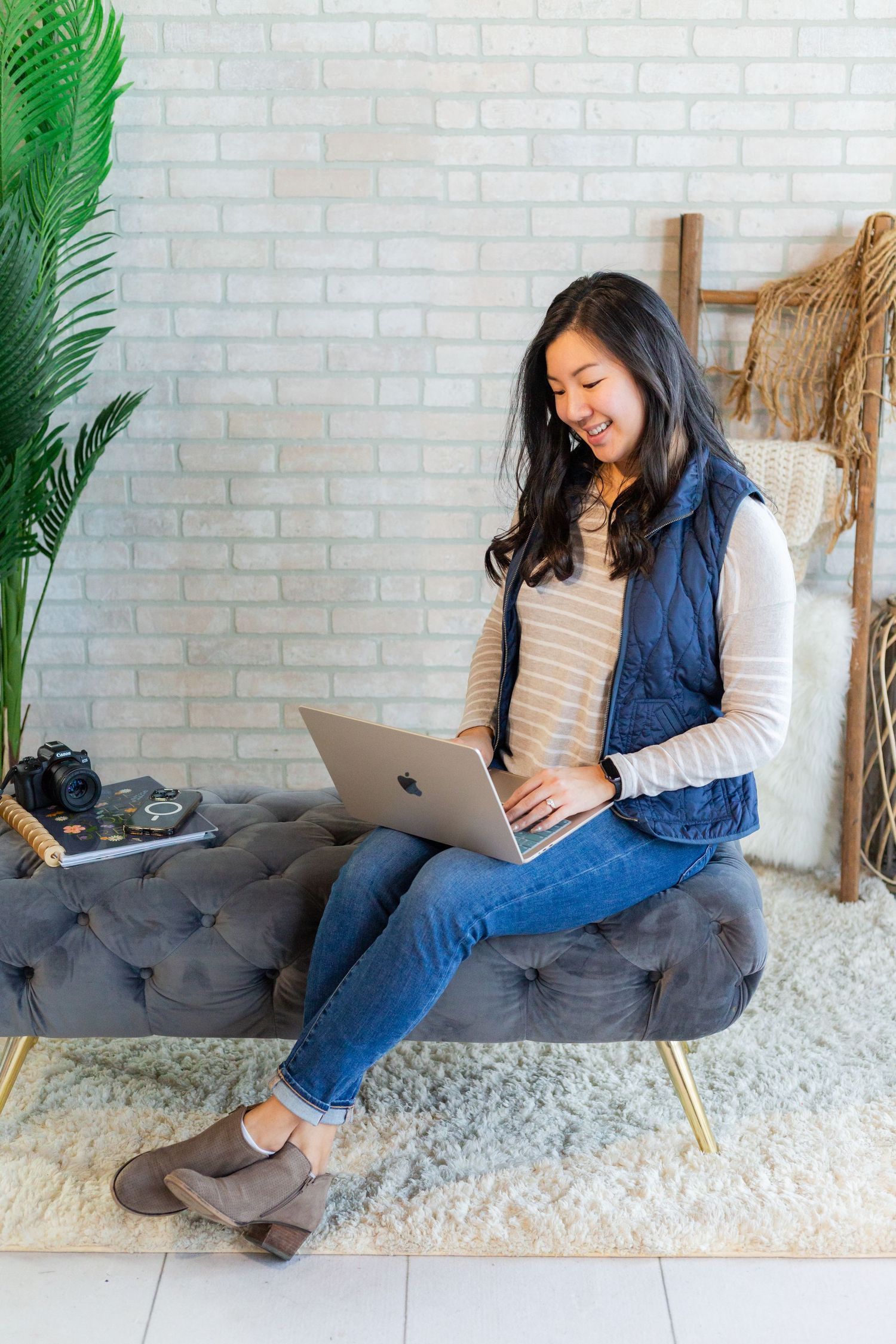 Smiling Asian woman sitting on a tufted bench using a MacBook laptop, wearing jeans and a navy vest.