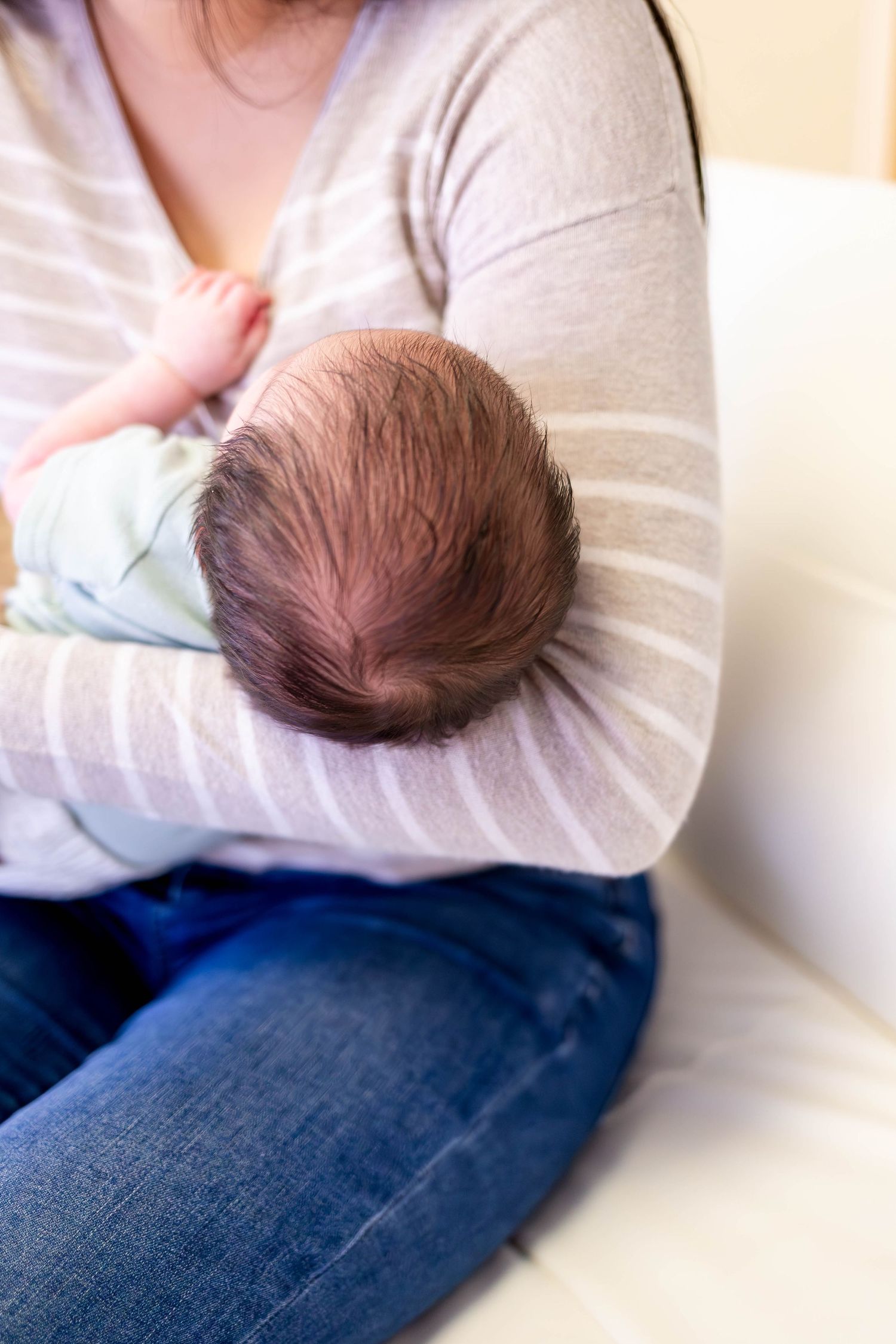 A newborns head rests on mom who is wearing a sweater and jeans while sitting on a couch in Central City photography studio.