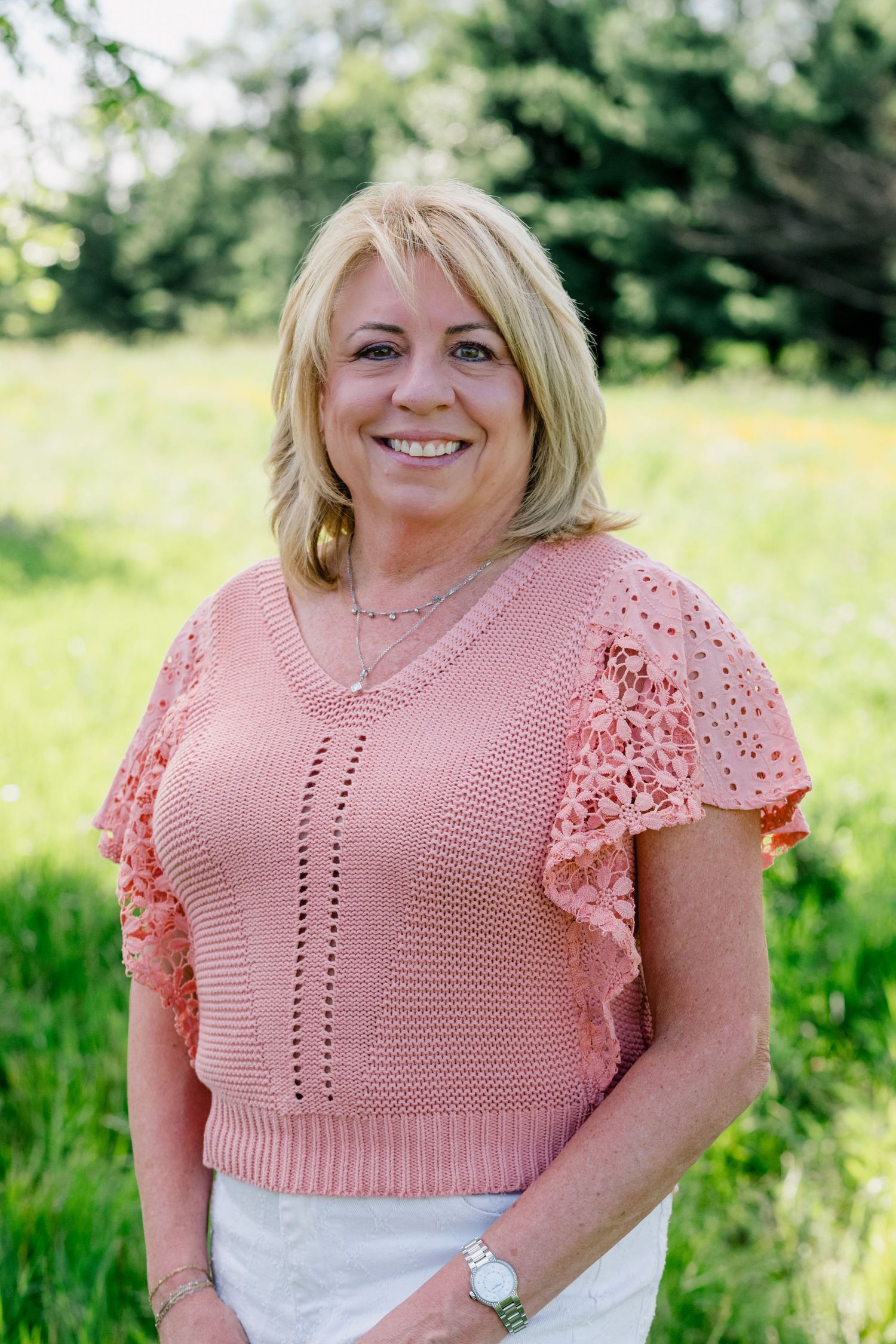 Smiling blonde woman in pink lace top posing outdoors in sunny green meadow.