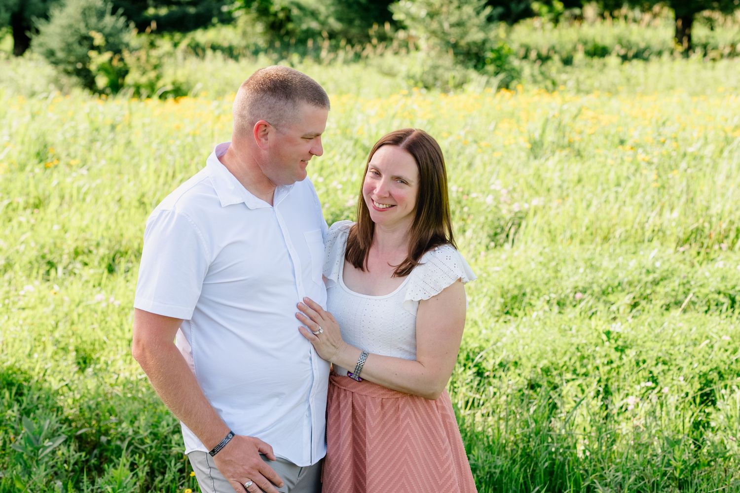 Couple smiling and embracing in a sunny green meadow with wildflowers, woman in pink skirt and man in white polo.