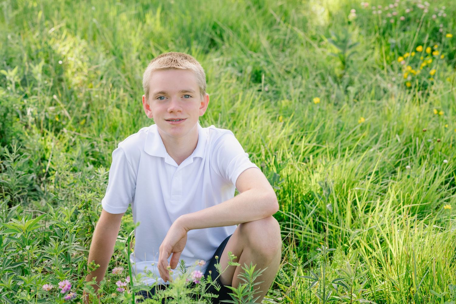 A smiling blond boy in a white polo shirt sits cross-legged in a lush green meadow with wildflowers.