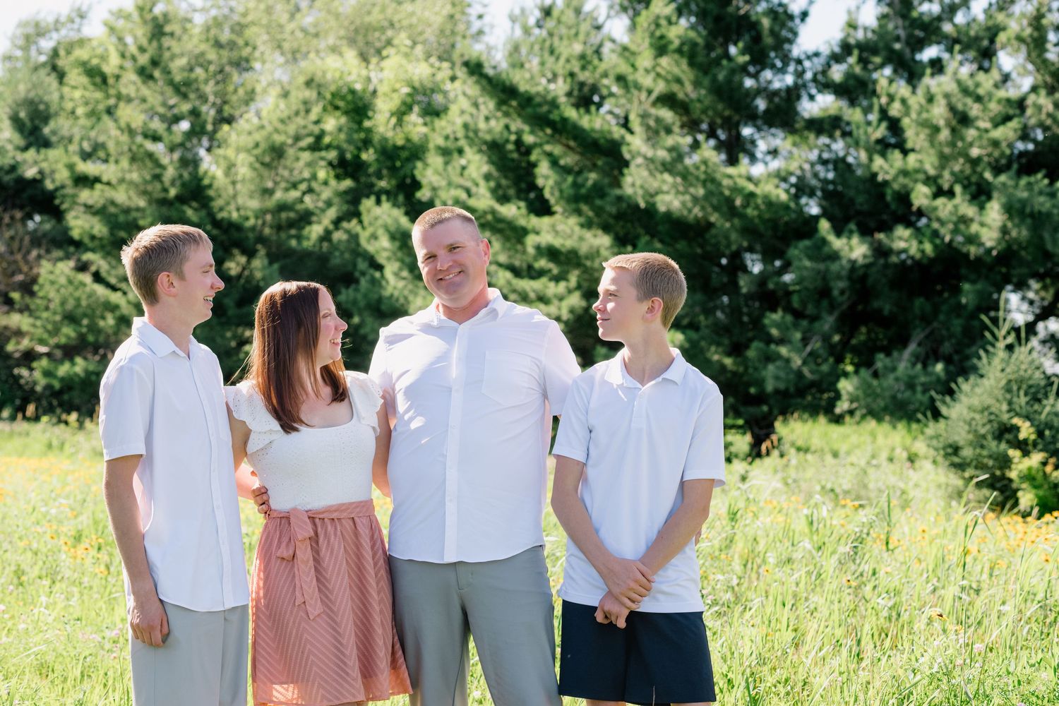Family of four smiling outdoors in a sunny green field, wearing casual white and gray outfits.