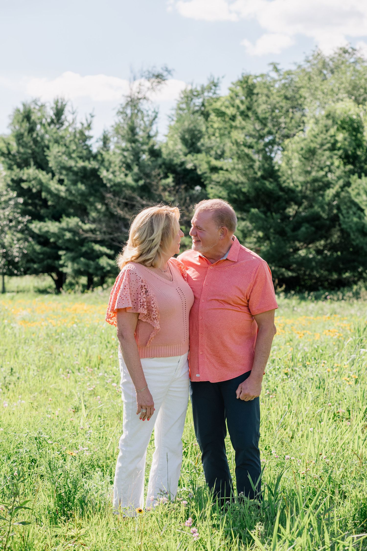 Couple in matching coral outfits smiling at each other in a sunny green meadow with wildflowers and trees.