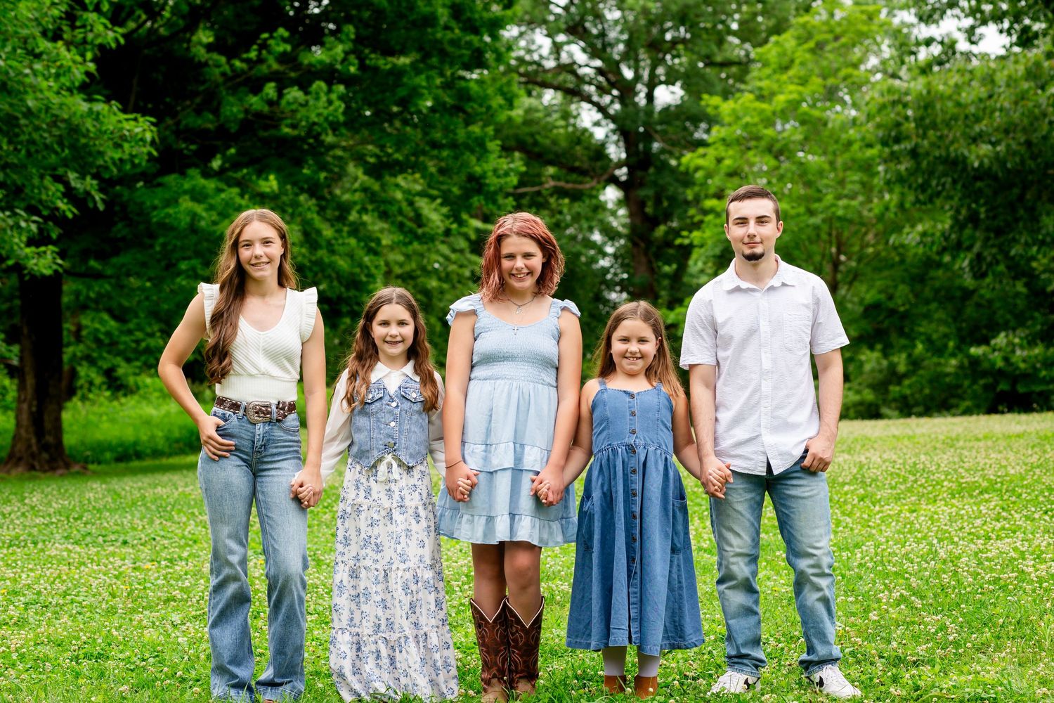 Family of five smiling together outdoors on green grass with trees in background on sunny day.
