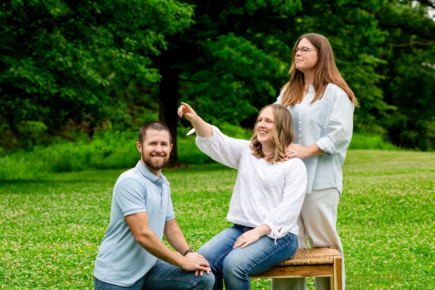Three adults laughing and posing together outdoors on a wooden bench in a green park.