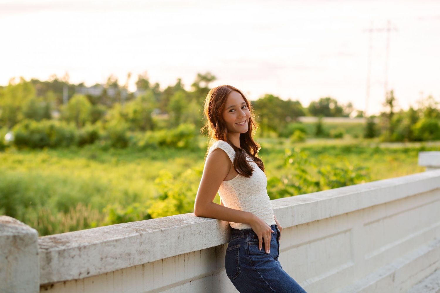 Young woman in white top and jeans leaning on a stone bridge railing at golden hour with lush green fields behind.