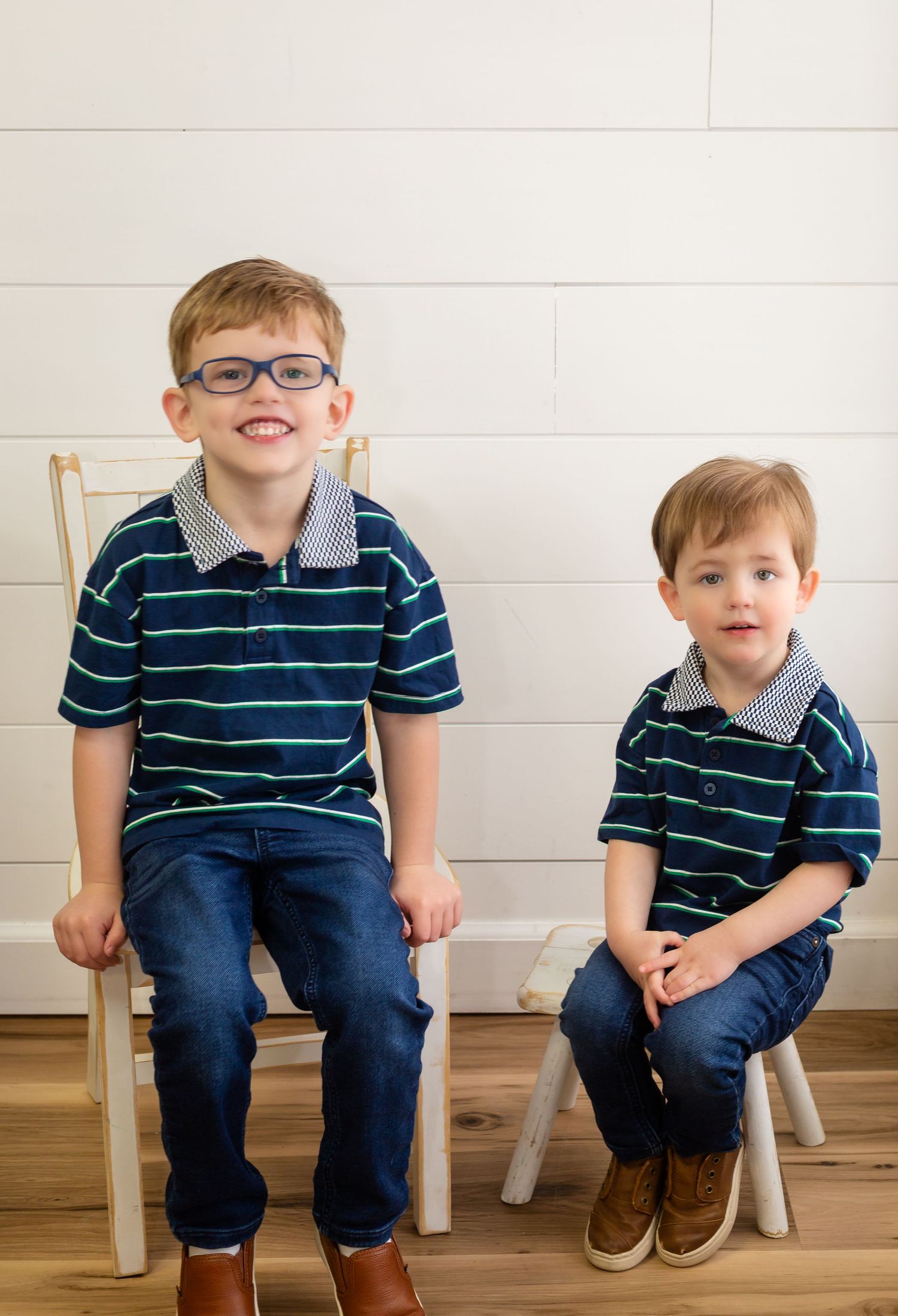 Two children in matching navy striped polo shirts and jeans sitting on white chairs against a white wall.