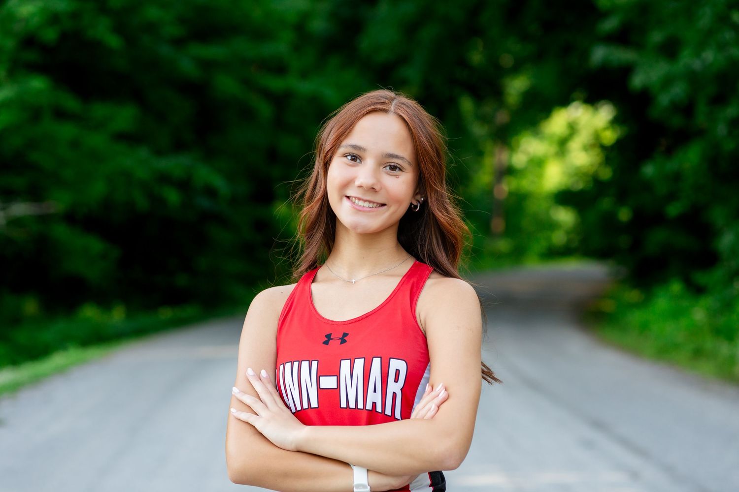 Smiling young woman in red Ann-Mar athletic tank top standing on a tree-lined road with arms crossed.