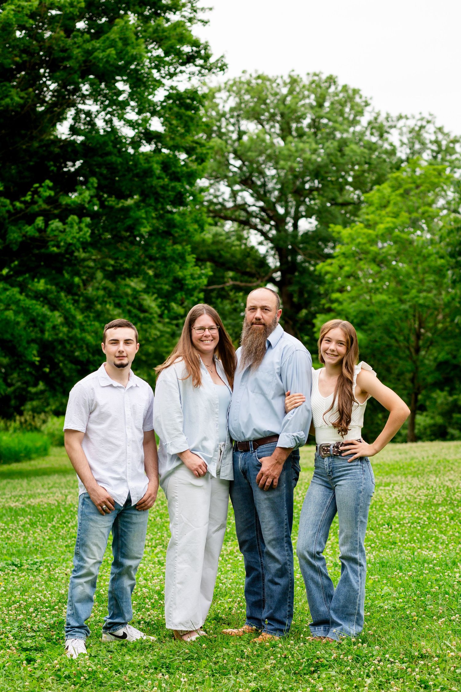 Family of four posing outdoors on green grass with trees, wearing casual blue and white outfits.