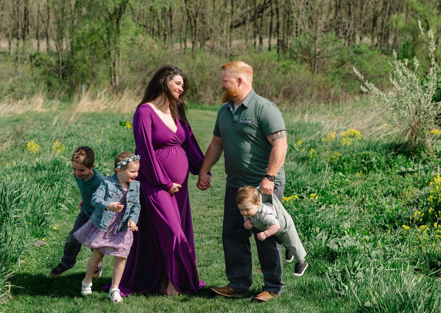 Pregnant woman in purple dress holds hands with husband while two young children play in a sunny green meadow.