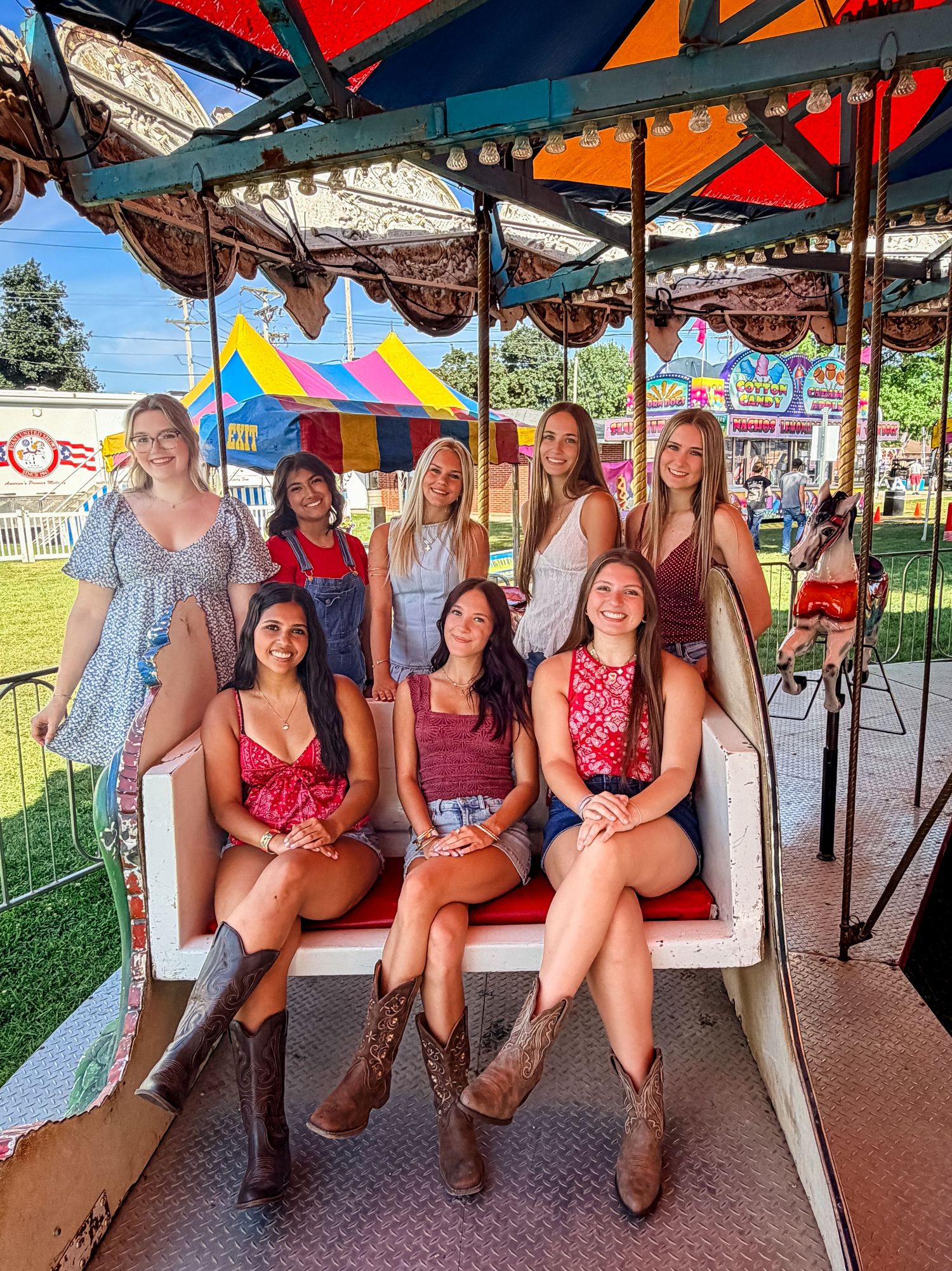 A group of eight young women posing together on a carnival ride at a summer fair, smiling and wearing casual summer outfits.