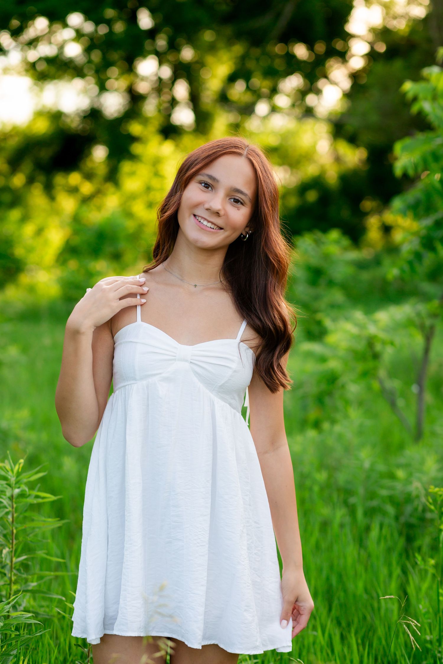 Young woman with red hair in white sundress smiling outdoors in lush green garden setting.