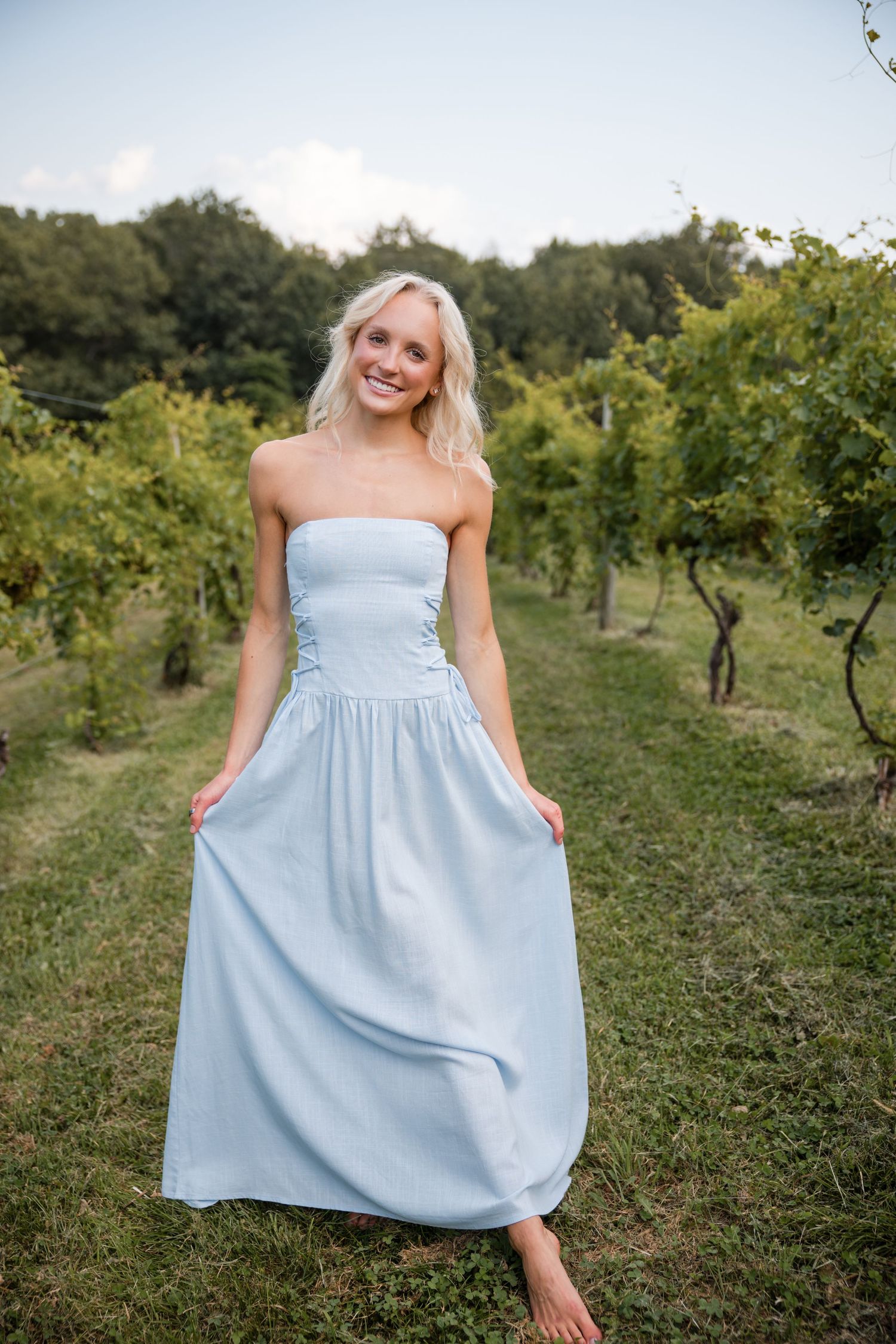 Smiling blonde woman in a light blue strapless maxi dress standing in a lush vineyard.