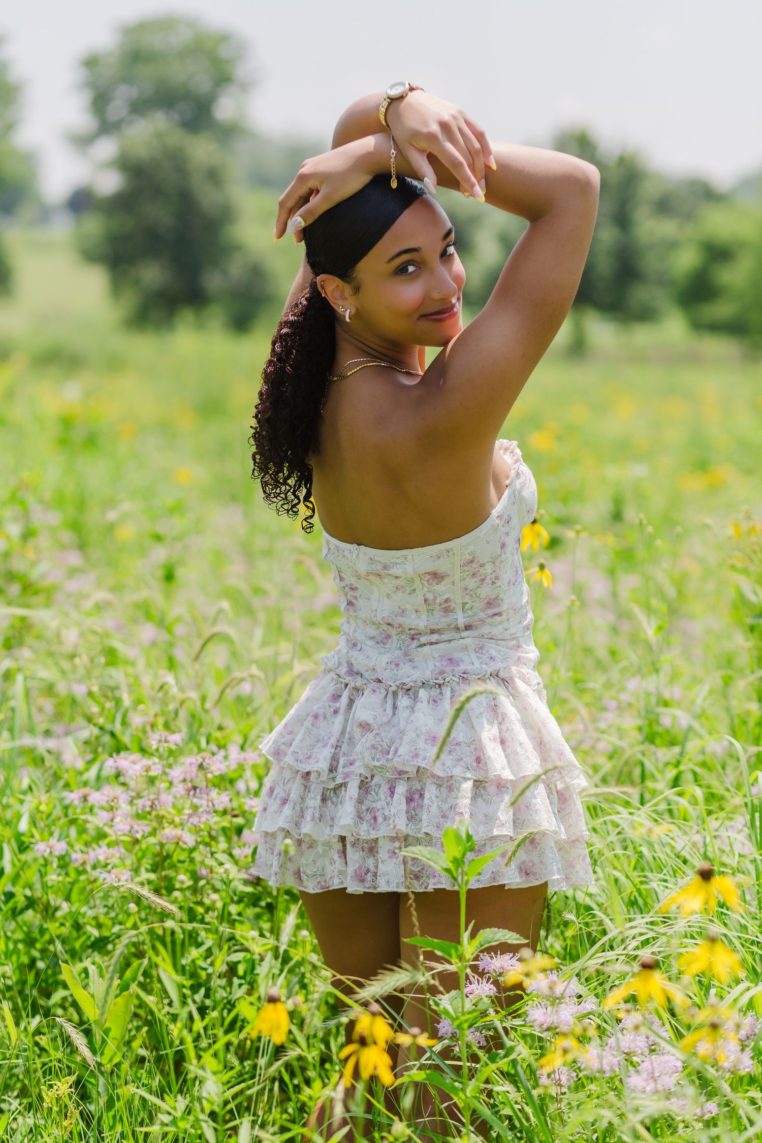 A high school senior girl in a white floral sundress poses gracefully in a meadow at Noelridge Park filled with yellow wildflowers.