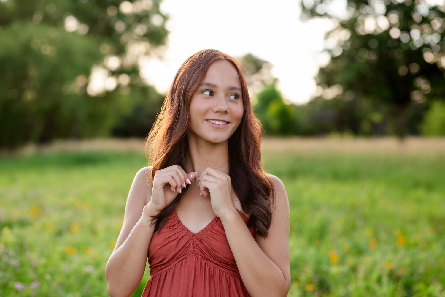 Young woman in a red dress smiling in a sunny green field with wildflowers in the background.