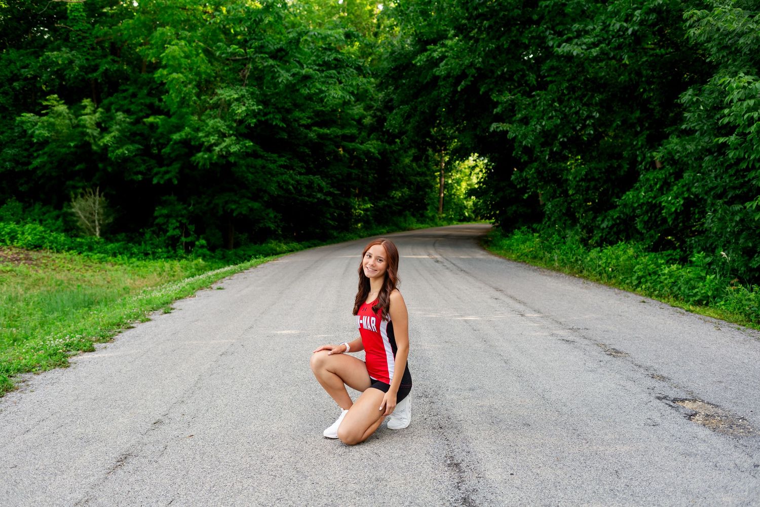 Young woman in red outfit crouching on a tree-lined road surrounded by lush green foliage.