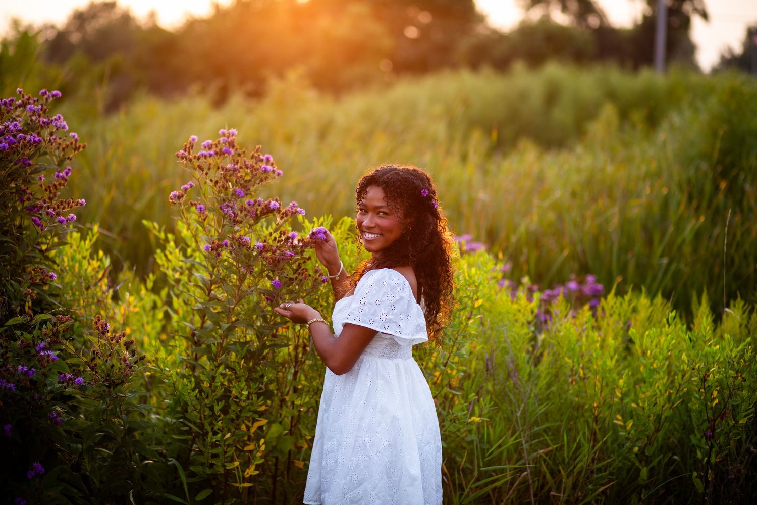 A person in a white dress stands among wildflowers in a meadow during golden hour sunset.