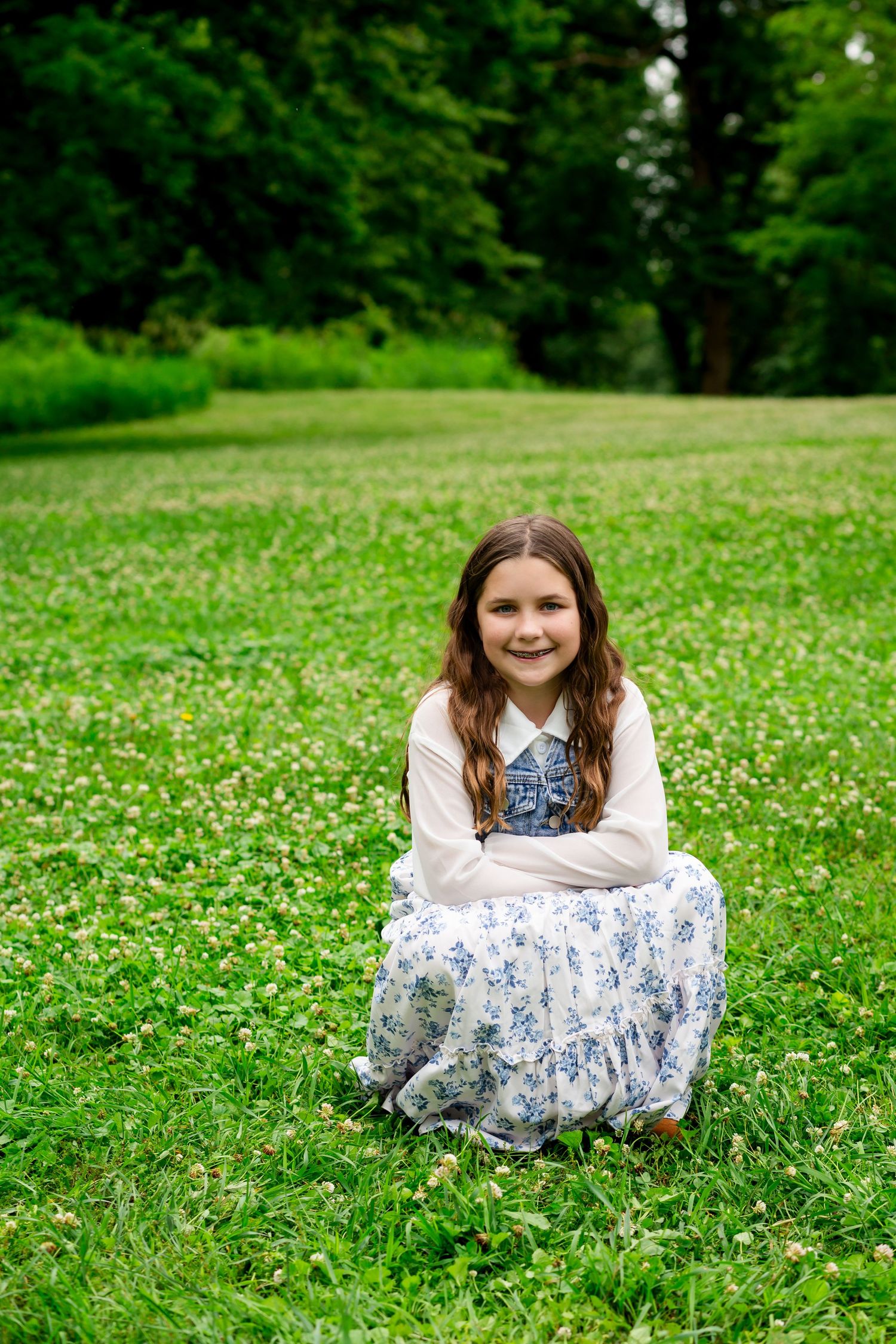 Young girl in floral dress sitting on green clover field, smiling at camera in lush park setting.