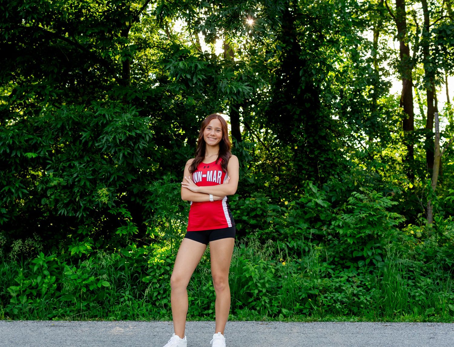 Young female athlete in red jersey and black shorts stands confidently with arms crossed on a path surrounded by lush green trees.