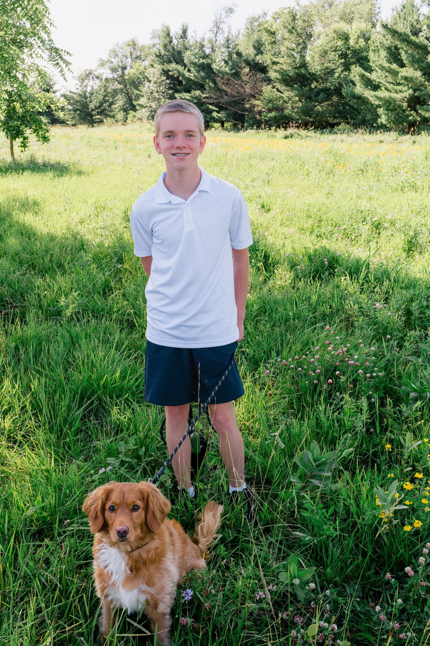 Smiling boy in white polo holding leash with golden dog in lush green field.
