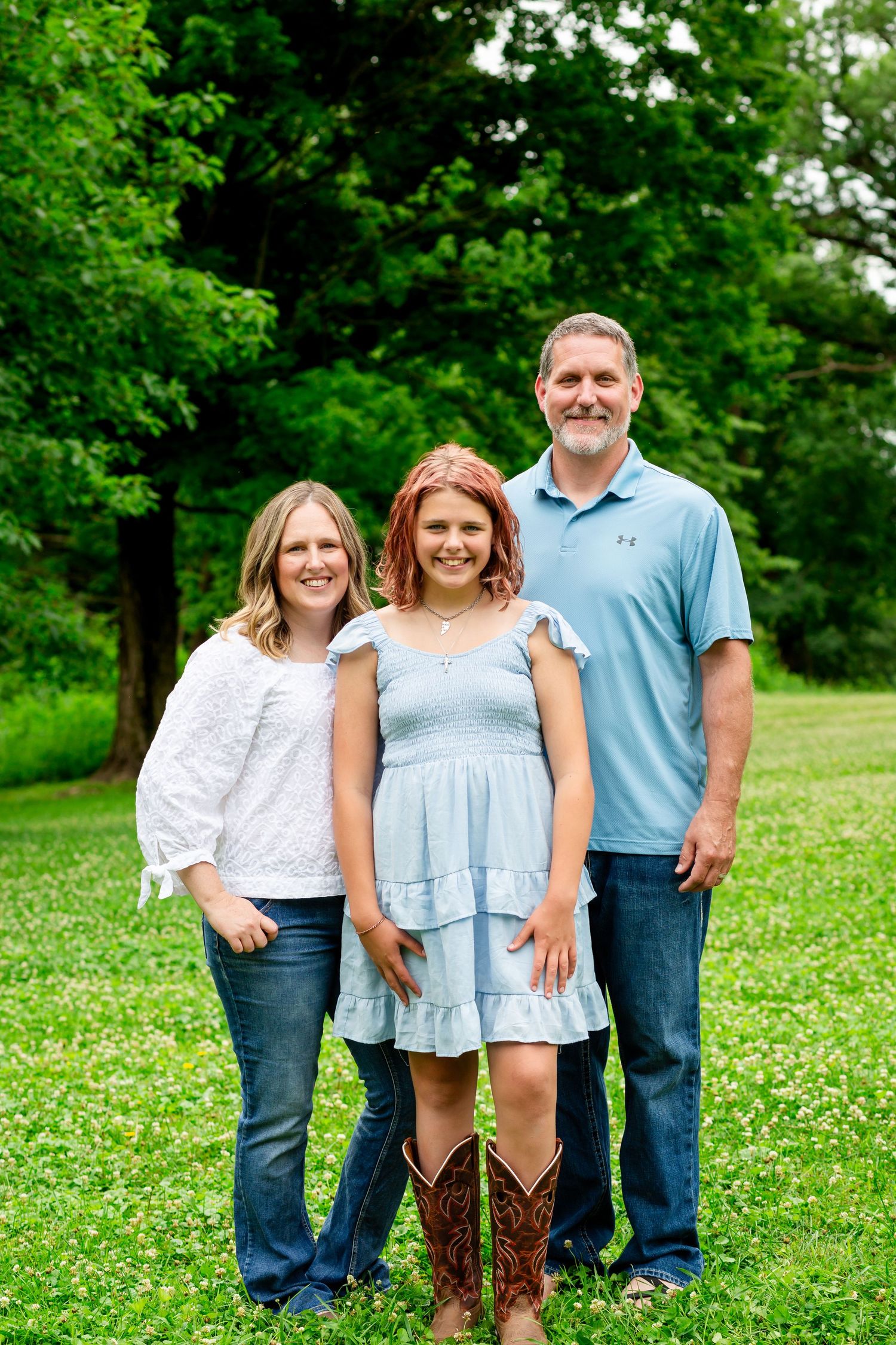 Family of three smiling outdoors, girl in blue dress with cowboy boots flanked by parents.