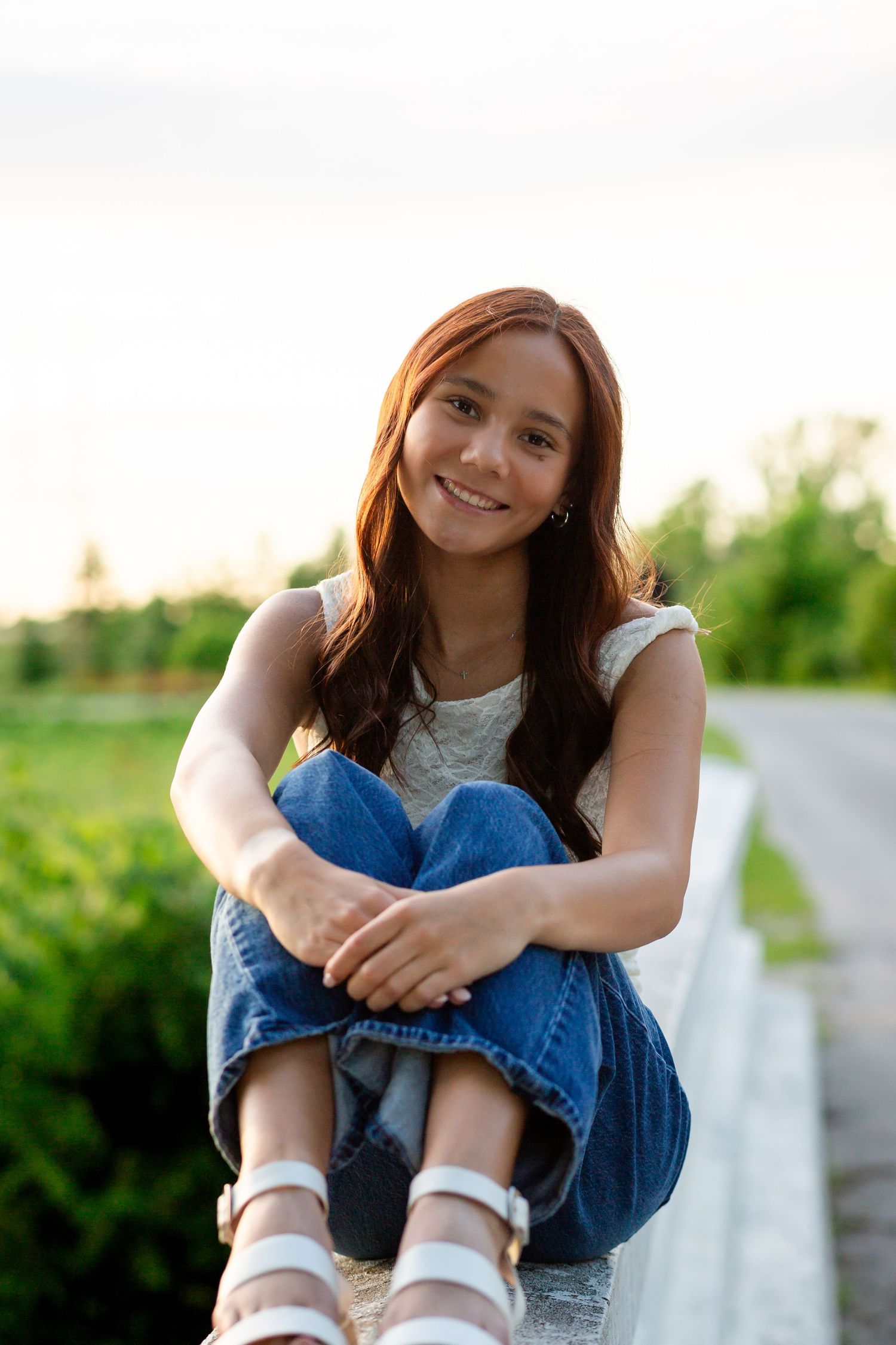 Young woman with red hair sitting outdoors in jeans and white top, smiling warmly at camera in natural green setting.