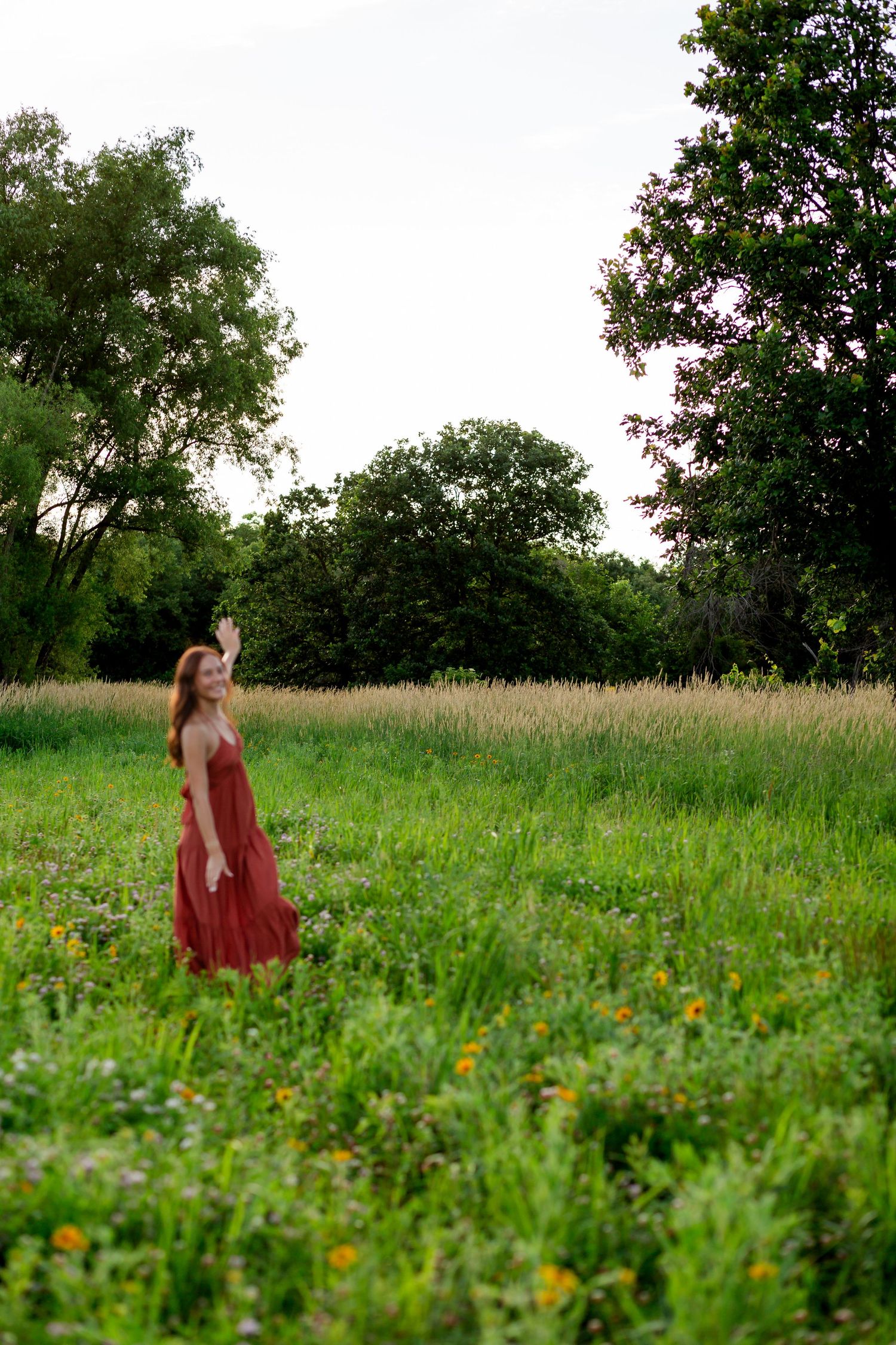 Woman in red dress standing in a lush green meadow surrounded by tall trees on a cloudy day.