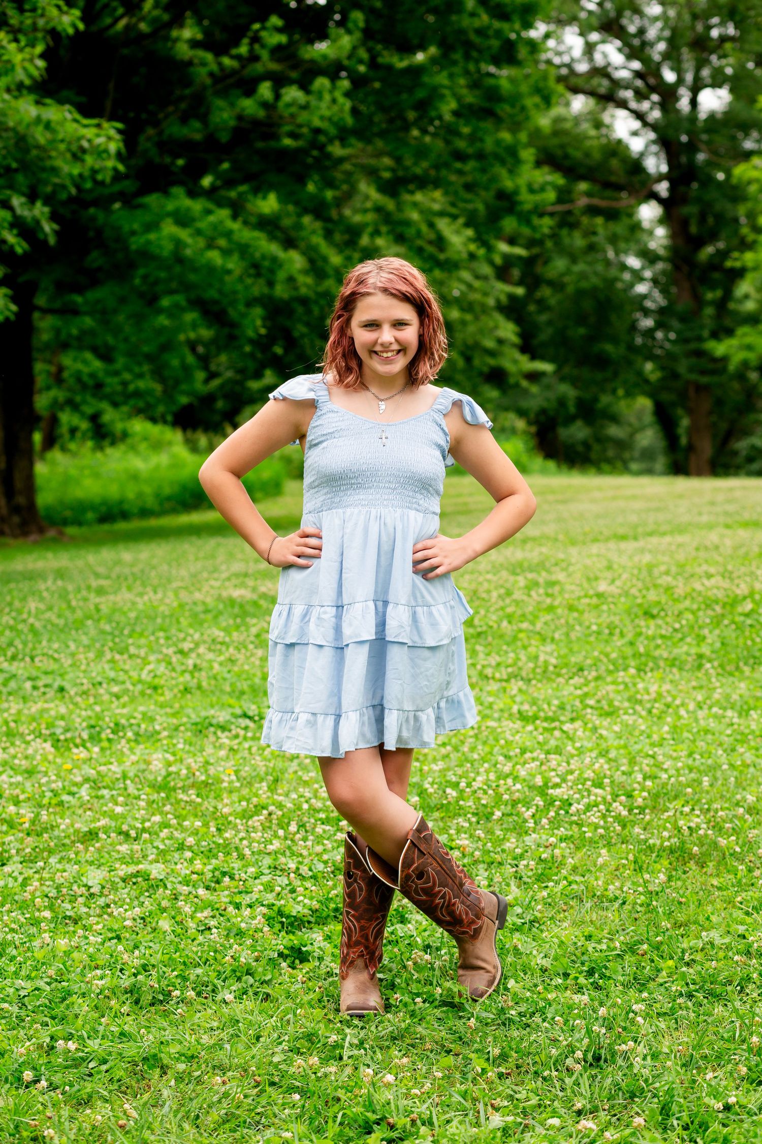 Young girl in light blue ruffled dress and cowboy boots posing in a green park with hands on hips.