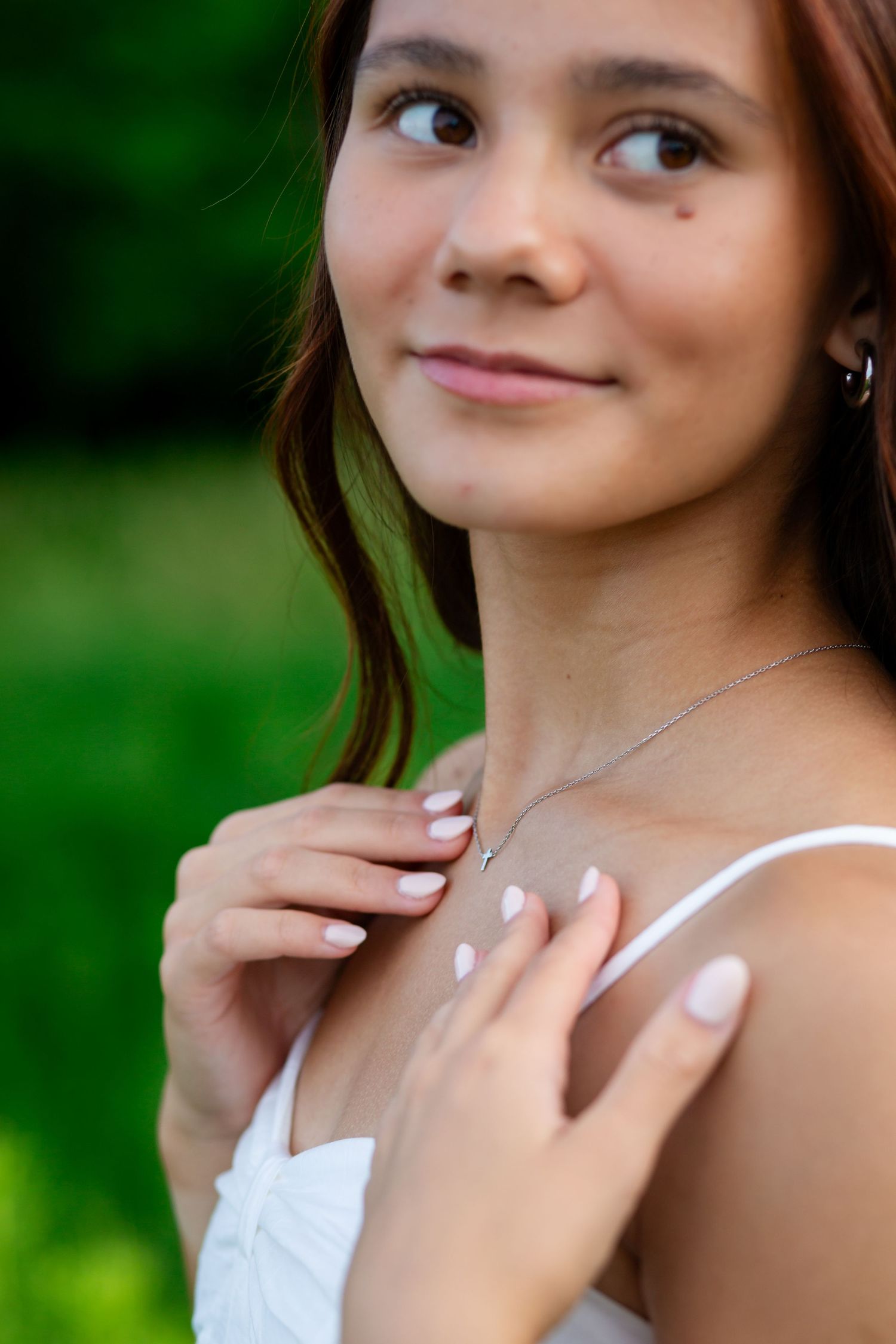 Young woman in white dress touching necklace, smiling softly outdoors with green bokeh background.