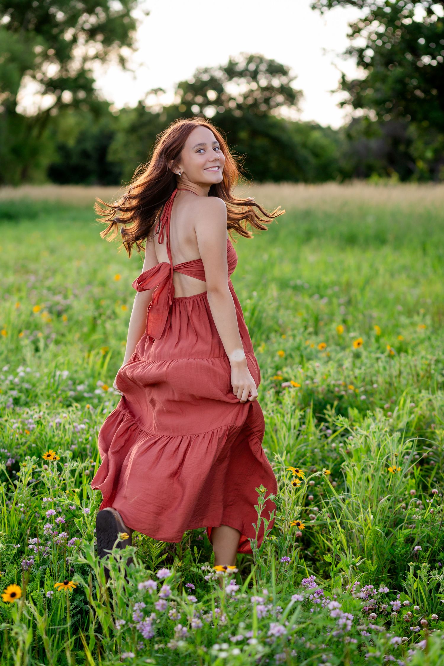 Redhead woman in rust maxi dress twirling through green wildflower field.