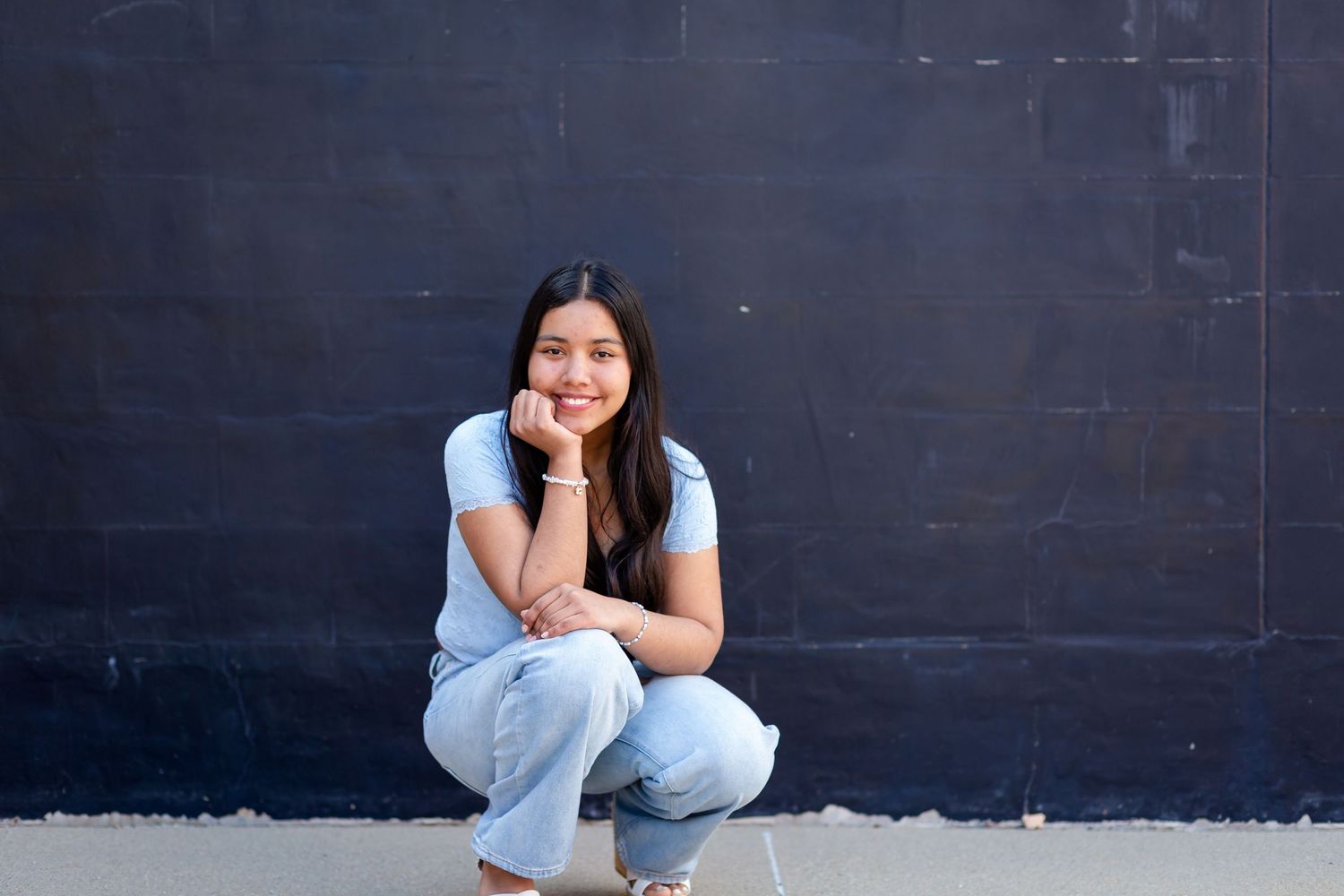 Young woman in light blue outfit crouching against dark wall, smiling in a casual outdoor portrait with her Marion senior photographer.