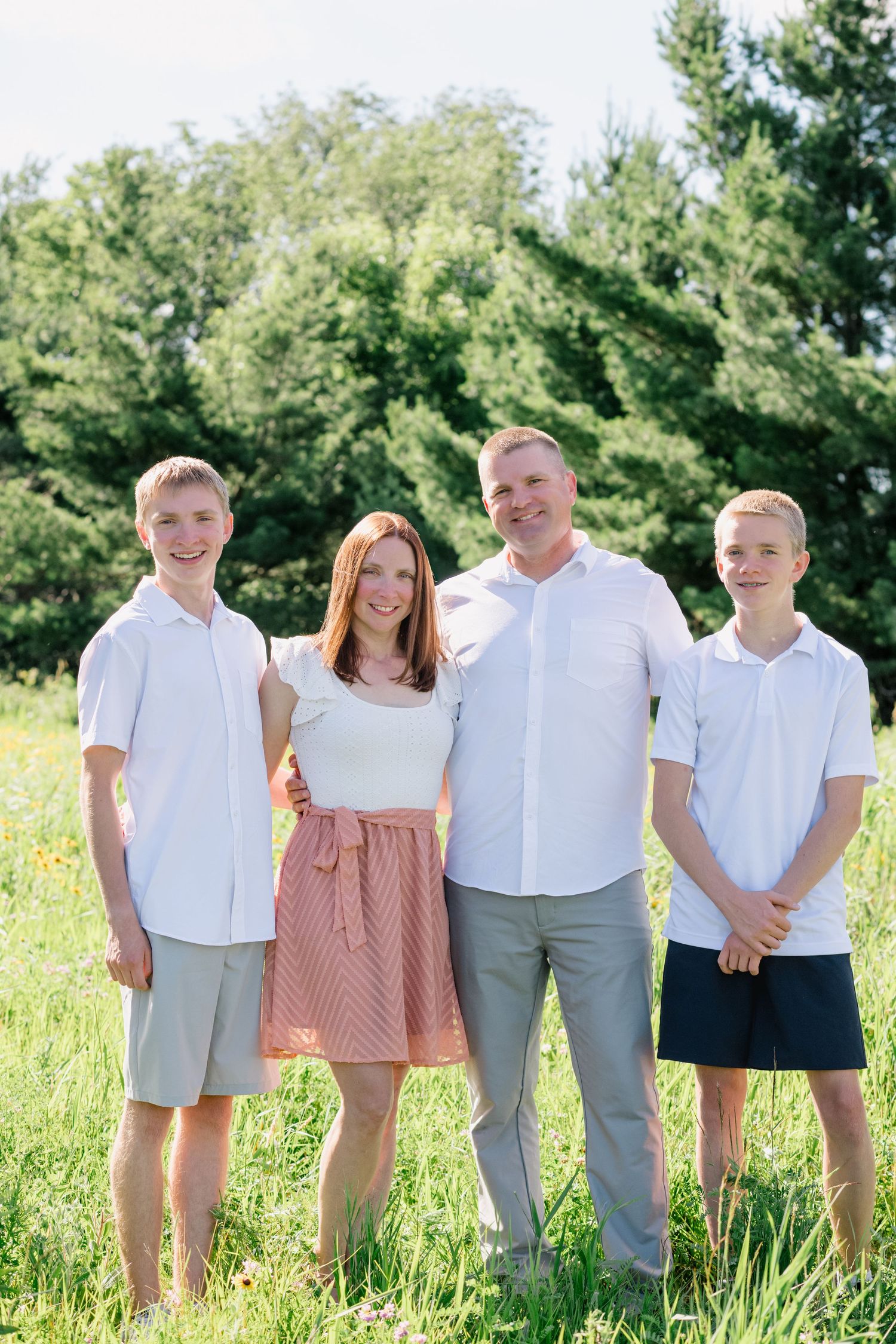 Family of four in white outfits smiling together in a sunny green field with wildflowers and trees.