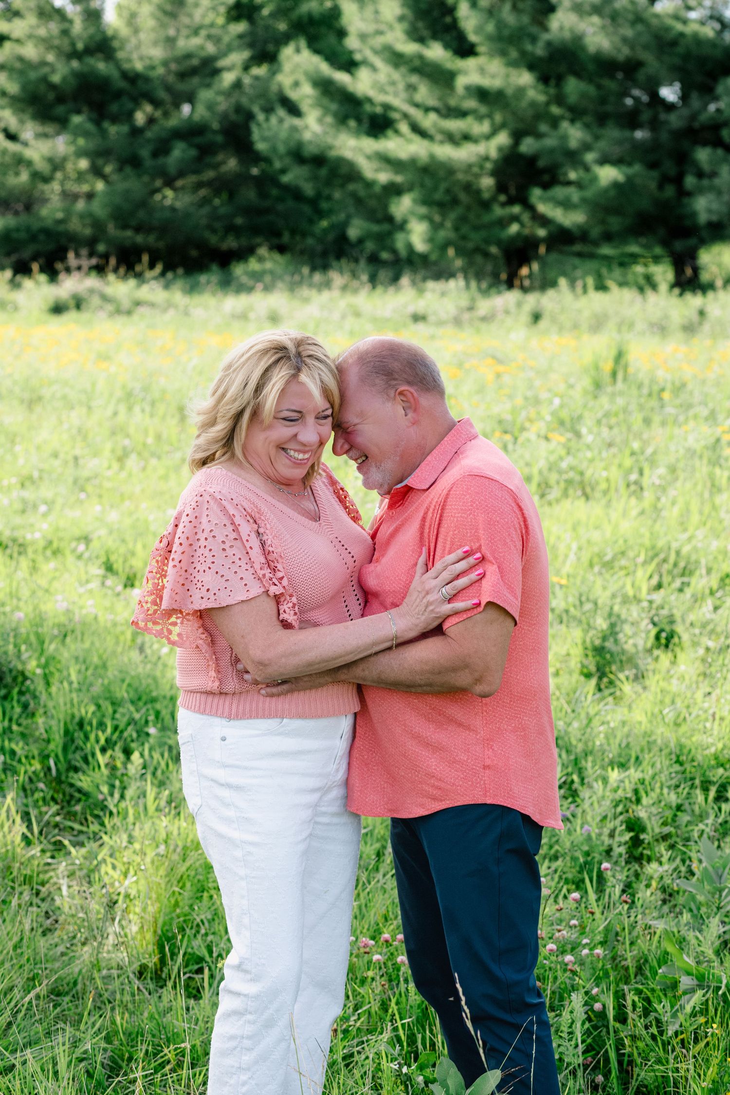 A smiling middle-aged couple embraces in a lush green meadow, wearing matching coral outfits on a sunny day.
