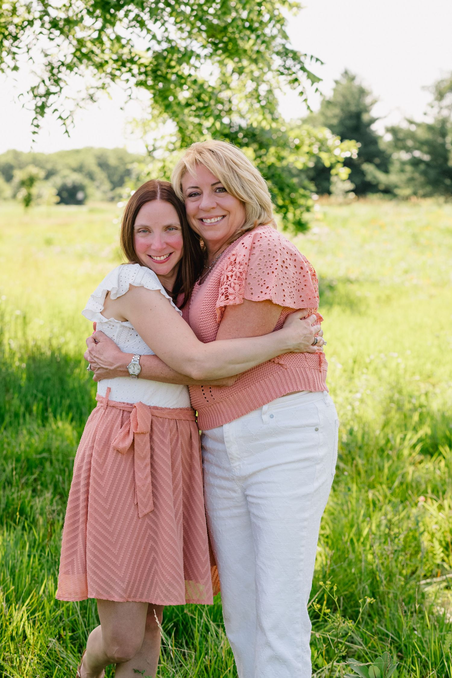Two women hugging and smiling outdoors in a sunny green meadow, wearing pink and white outfits.