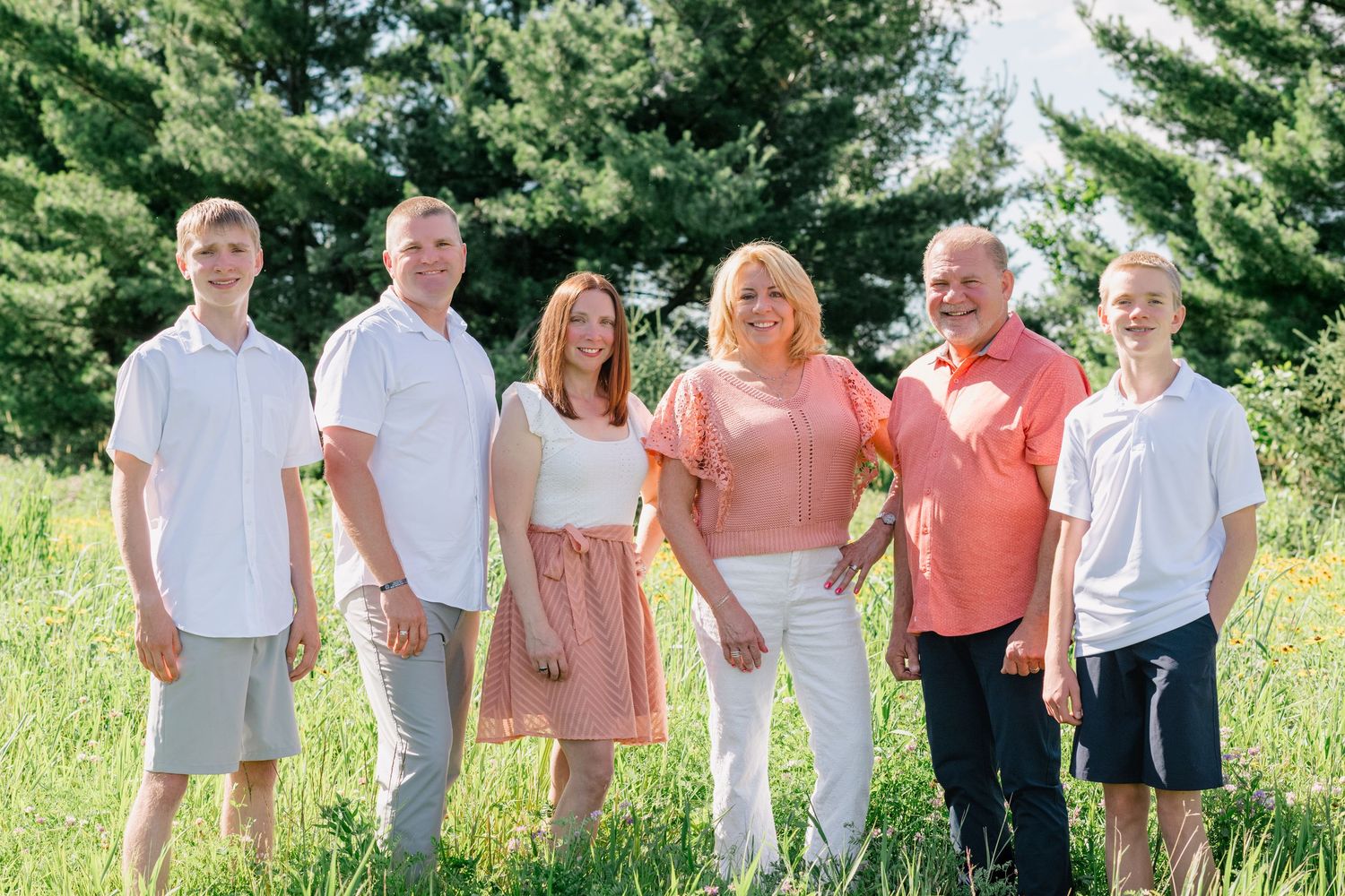 A happy family of six poses outdoors in a green meadow with tall trees in the background on a sunny day.
